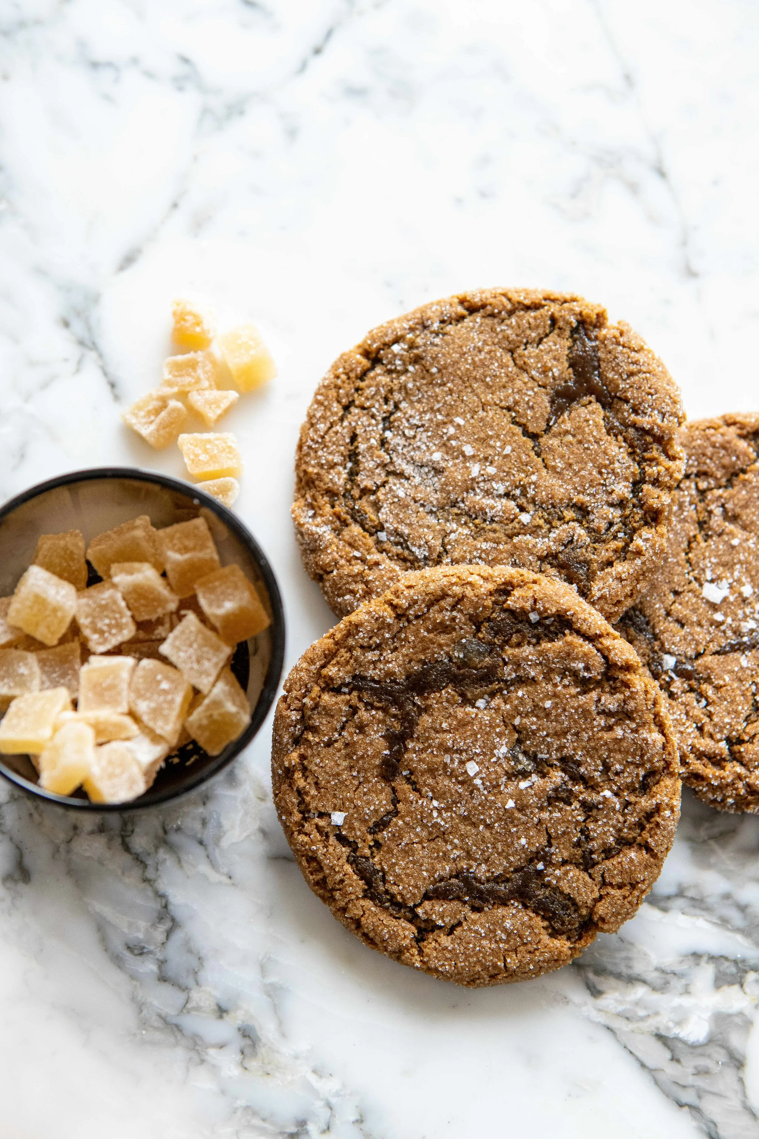 Three chocolate chip cookies dusted with powdered sugar, with a small bowl of white sugar cubes beside them on a white marble surface.