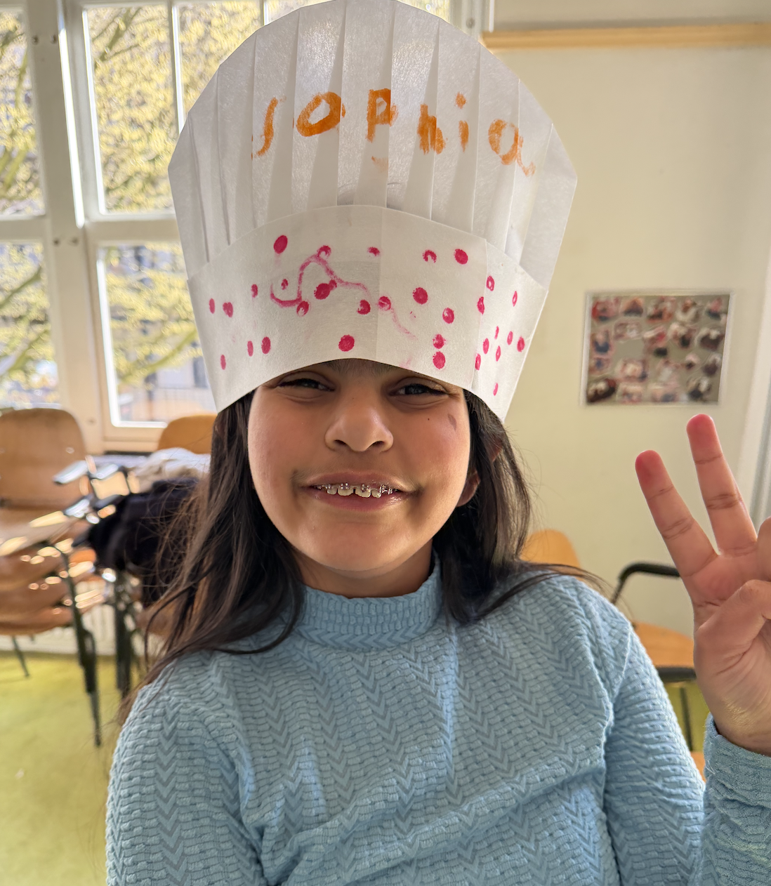 Child wearing a paper chef's hat decorated with pink dots and the name 'Sophia' written in orange, smiling and making a peace sign indoors near a window.