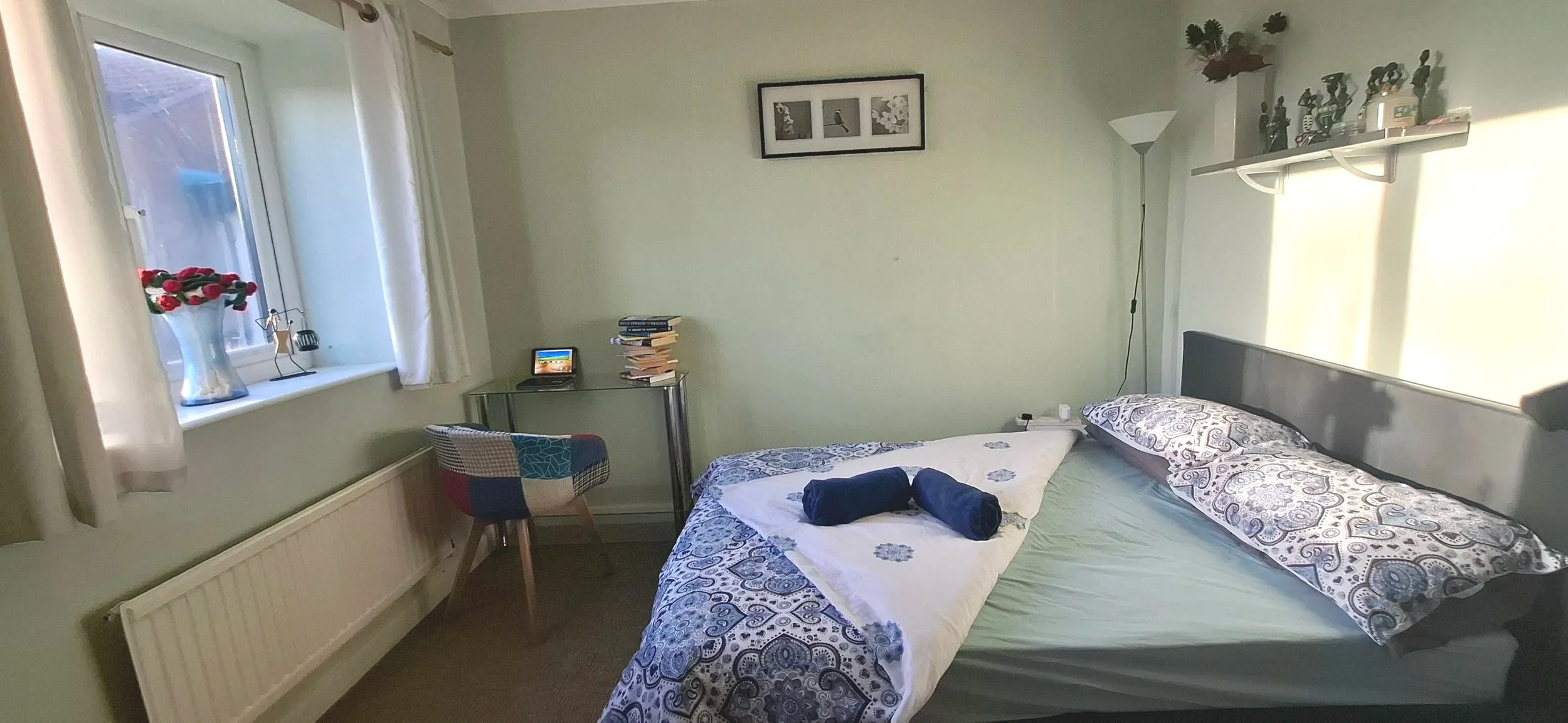 A double bed with light streaming in via the window opposite. to the left is a glass table and a bucket seat chair.