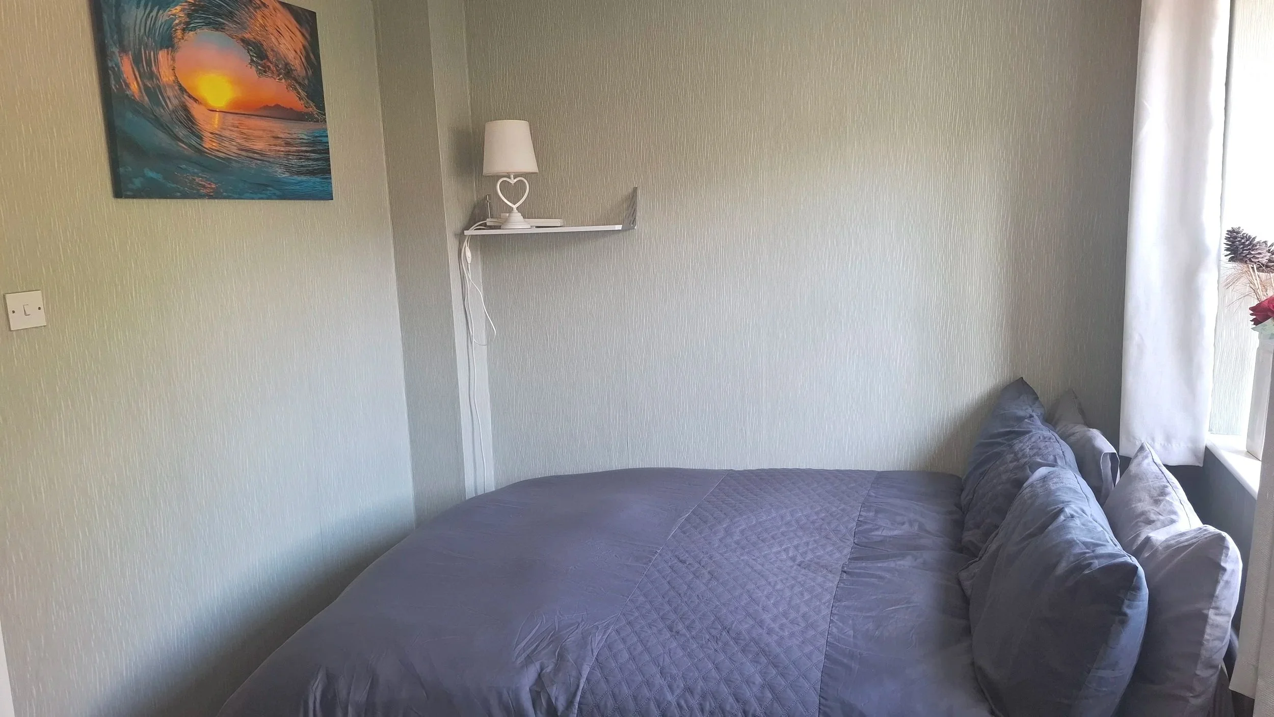 A bedroom with a gray quilted bedspread, a gray pillow, a white side table holding a glass pitcher, a white pitcher, and glasses, and a colorful ocean wave and sunset painting on the wall.