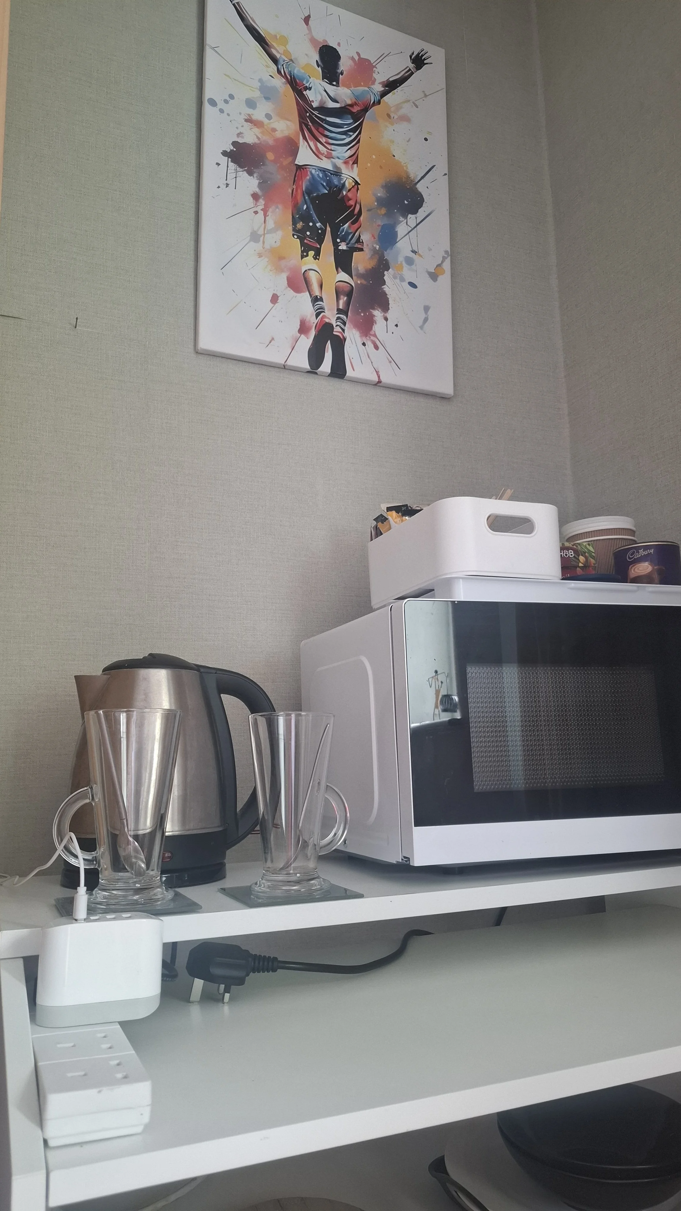 A white kitchen counter with a microwave, a basket of assorted snacks and tea, two ceramic mugs with pink and grey patterns, a stainless steel electric kettle, and a power strip.