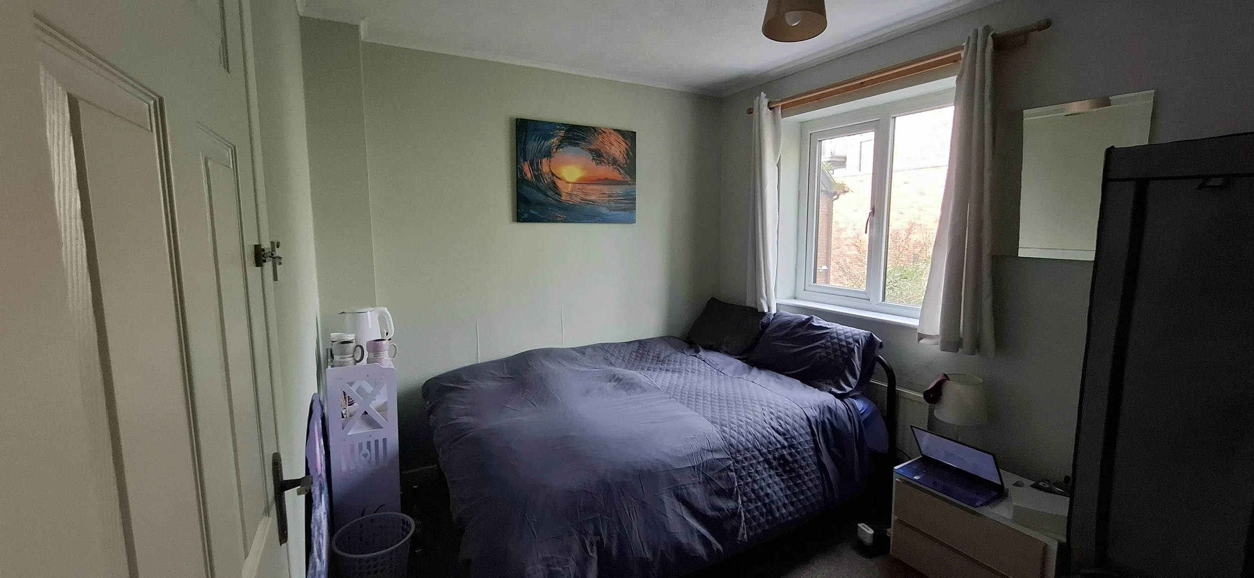 A bedroom with a gray quilted bedspread, a gray pillow, a white side table holding a glass pitcher, a white pitcher, and glasses, and a colorful ocean wave and sunset painting on the wall.