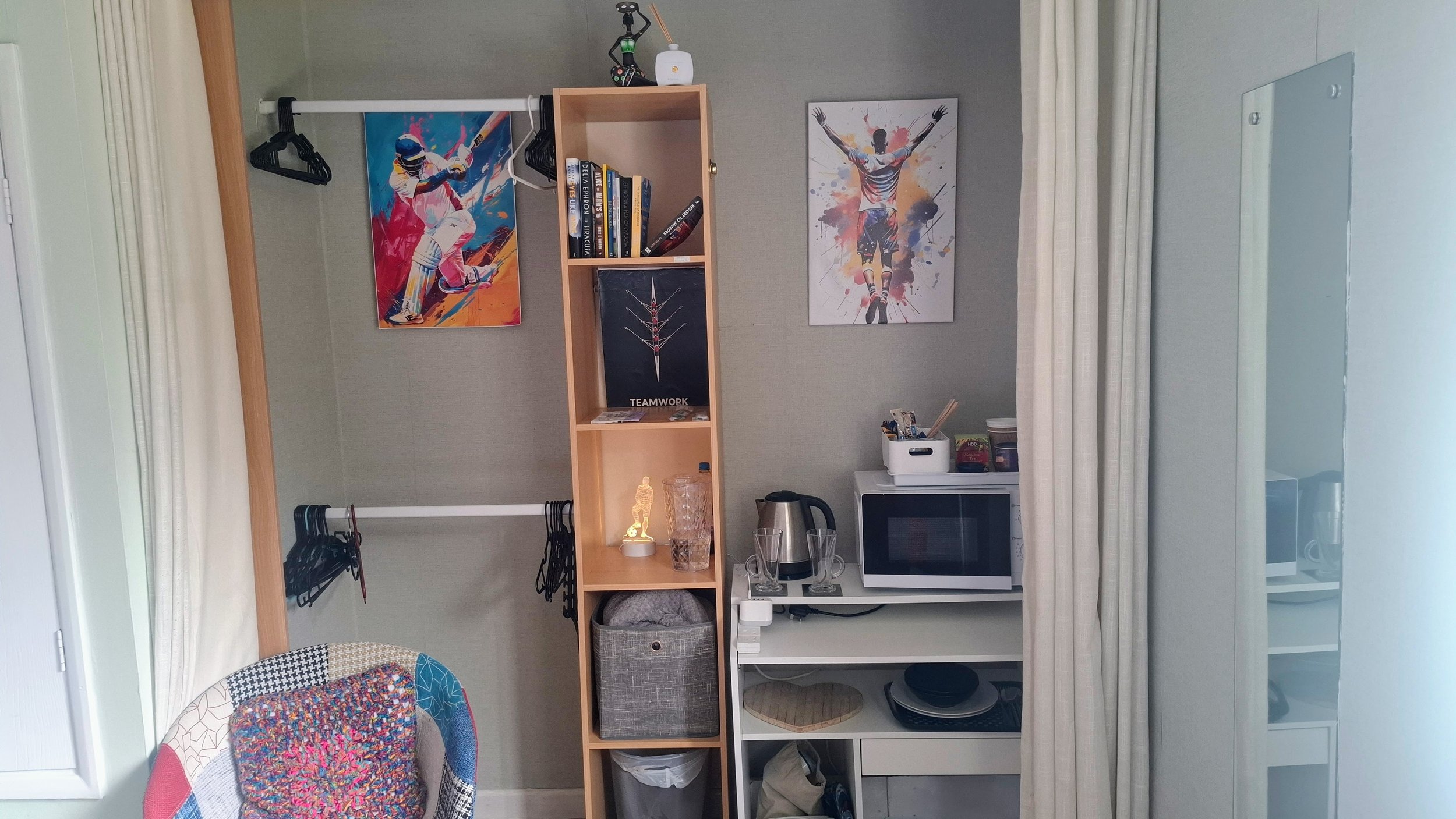 A cozy room space with beige carpeting, cream-colored curtains, a small multicolored rug, a tall wooden shelving unit filled with various objects including glassware, a gray towel hanging on the shelf, a small white cabinet with a microwave on top, and a framed picture of a tiger on the wall.