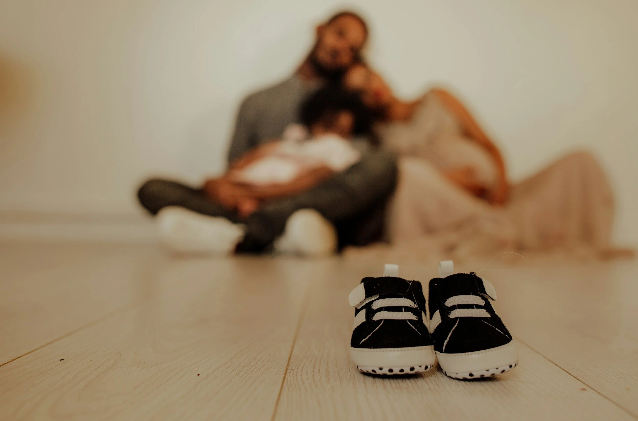 Pair of small black and white baby shoes on a wooden floor with blurred people sitting on the floor in the background.