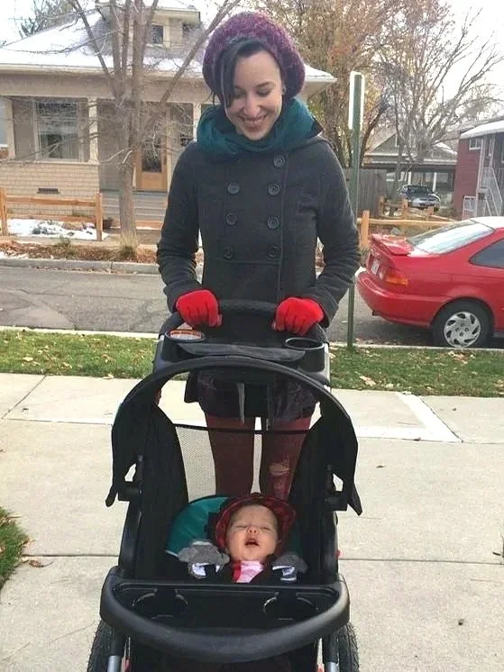 A woman smiling and pushing a baby in a stroller on a sidewalk in a residential neighborhood during fall or winter weather.