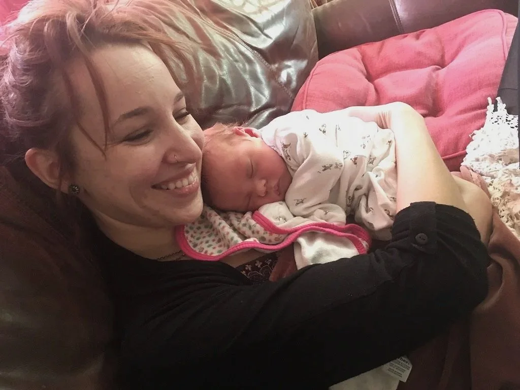 A woman with red hair smiling and holding a sleeping baby on her shoulder on a brown leather couch, with a pink stuffed animal nearby.
