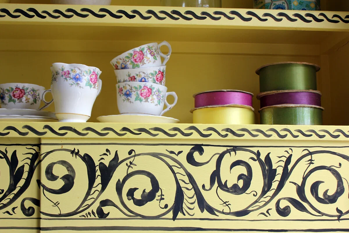 Yellow cabinet with black decorative scrollwork, holding floral teacups, saucers, and colorful round tins.