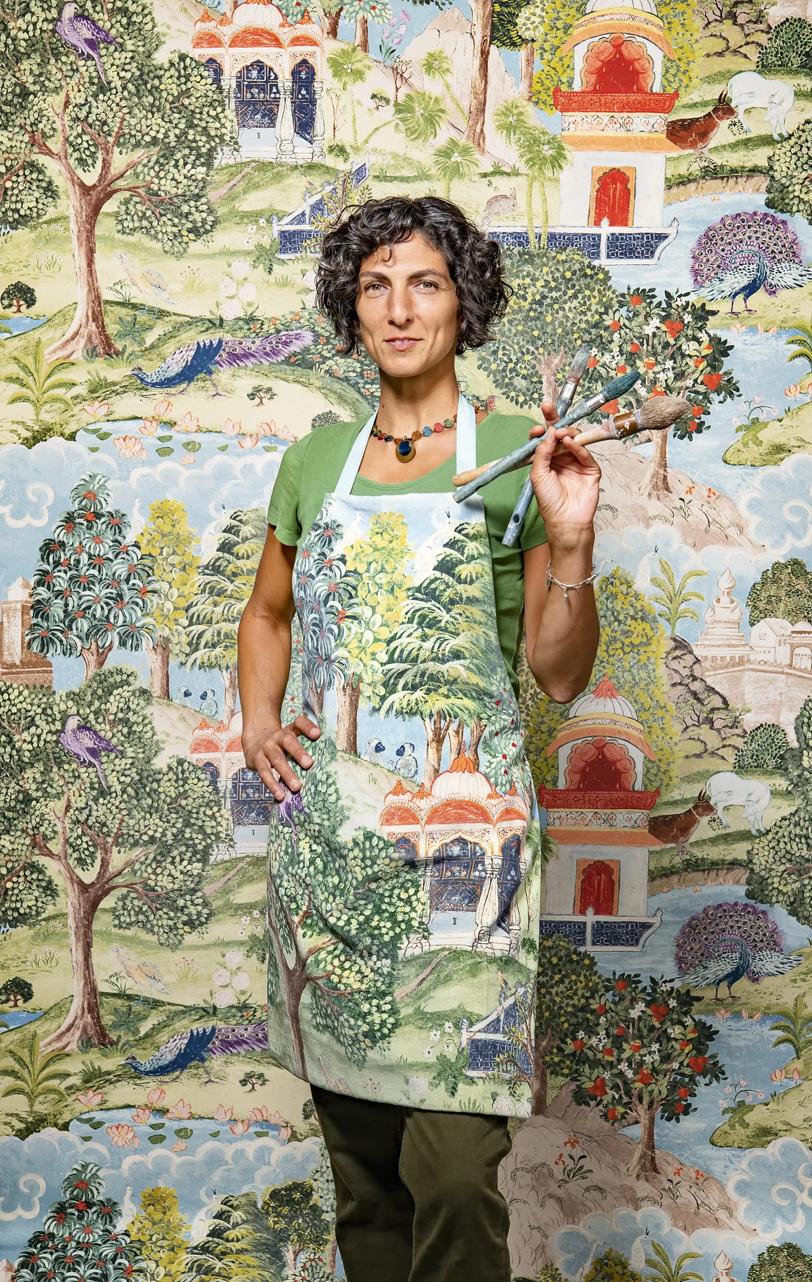 A woman standing in front of a colorful mural wearing an apron with nature scenery. She holds three paintbrushes and looks at the camera.