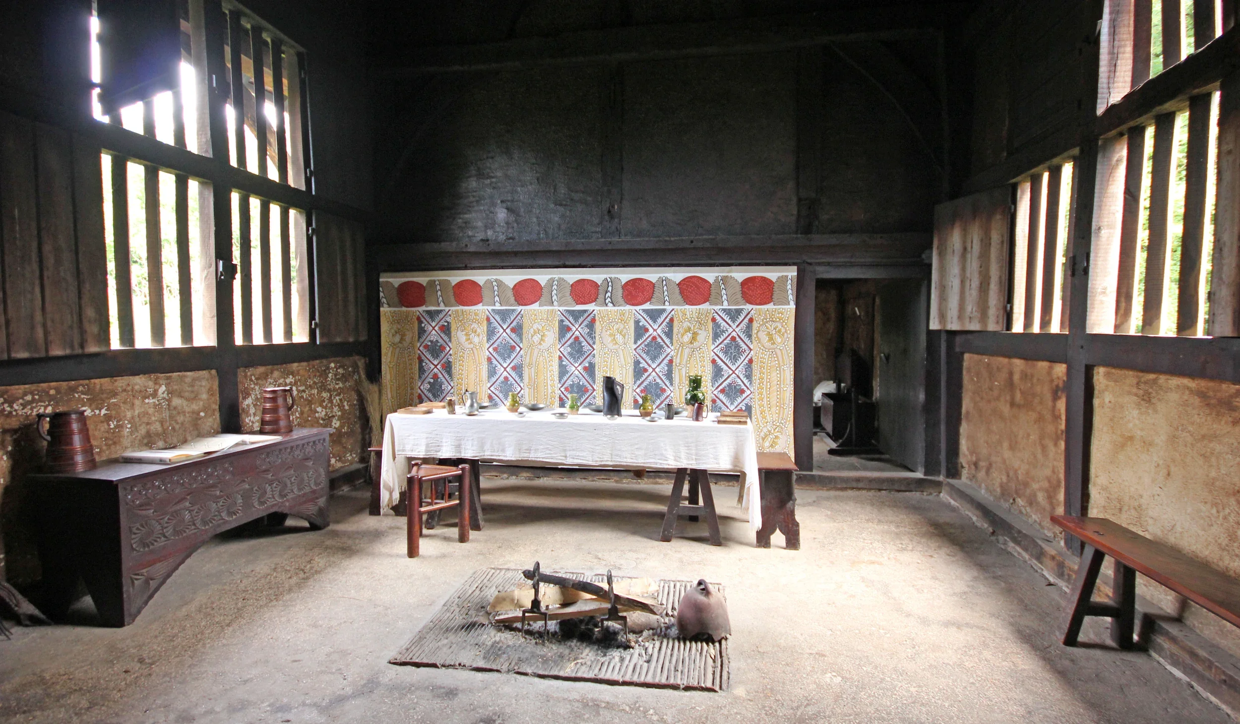Interior of a rustic wooden building with dark walls and wooden slats on the windows. There is a white tablecloth-covered table in the center with various decorative items and tableware. A folding chair is placed at the near end of the table. The back wall has a colorful patterned fabric or mural, and there are wooden benches on either side of the room. Sunlight filters through the window slats, illuminating parts of the room.