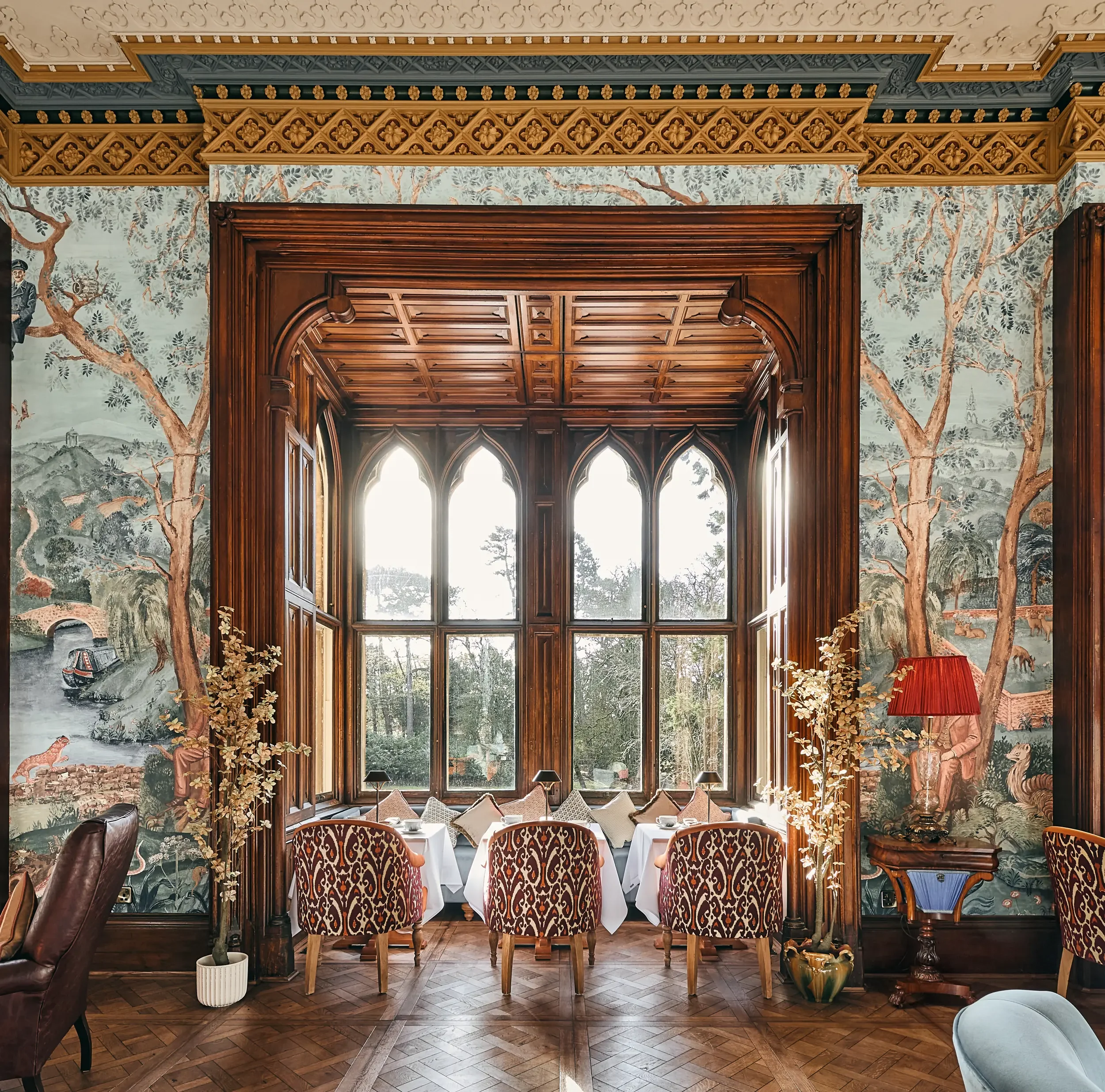 A luxurious dining room with grand wooden architecture, large arched windows, and ornate painted wallpaper depicting trees and countryside scenery. Stylish chairs surround a dining table set with cups and plates, and decorative plants and furniture complete the elegant decor.