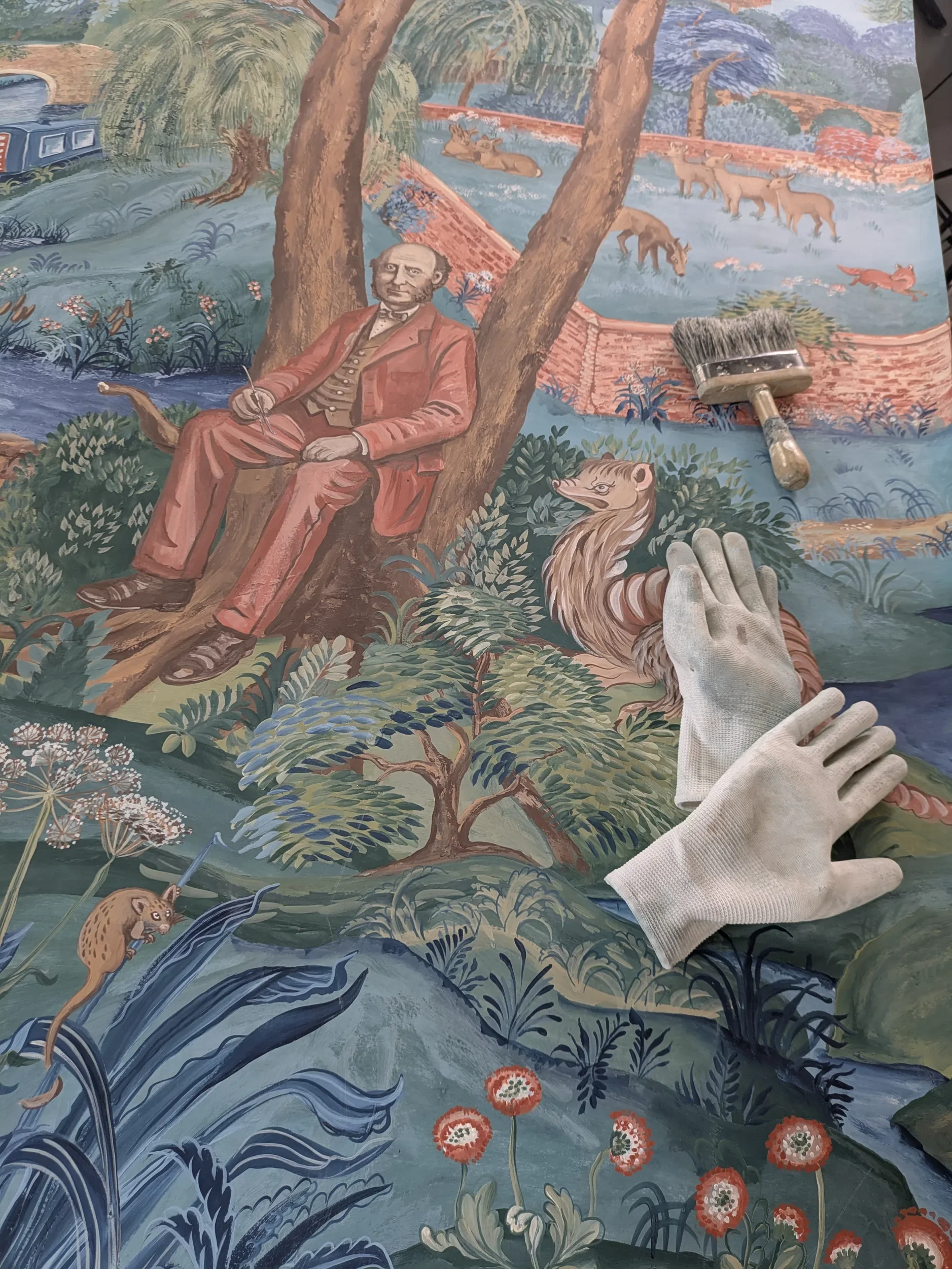 A detailed mural painting depicting a natural landscape scene with trees, flowing water, a train on tracks in the background, and various animals including a lion, a hyena, a pig, and deer, with a painter's brush and gloves in the foreground.