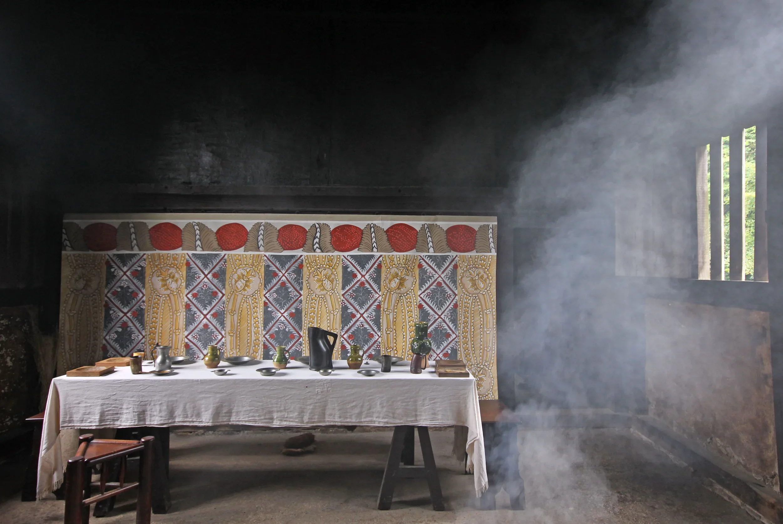 Interior of a rustic room with a large hanging tapestry featuring a floral pattern and warm colors. A dining table with a white tablecloth and various pottery items is in front of the tapestry. Smoke or mist hangs in the air, and a window with vertical slats letting in light is on the right side of the room.