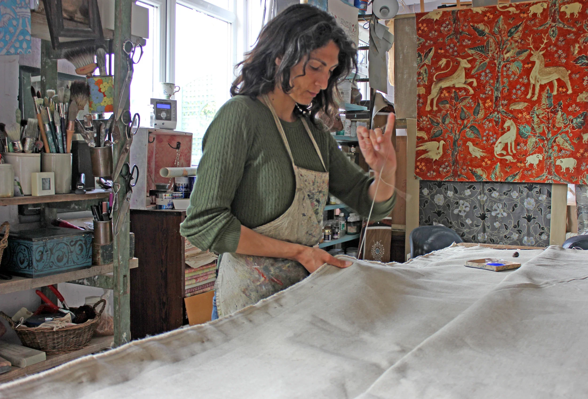 A woman with dark curly hair, wearing a green sweater and an apron, working on a fabric project in a light-filled craft room with colorful textiles on the wall, shelves with art supplies, and a table covered with fabric.