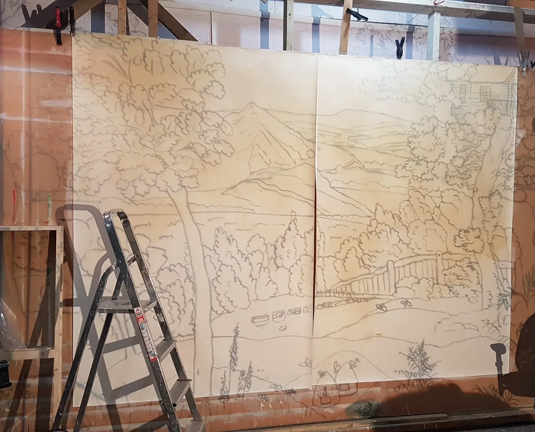 A large wooden panel with a detailed outline of a landscape scene, including trees, mountains, a house, and a fence, is secured to a wall with clamps; a ladder is positioned in front of it.
