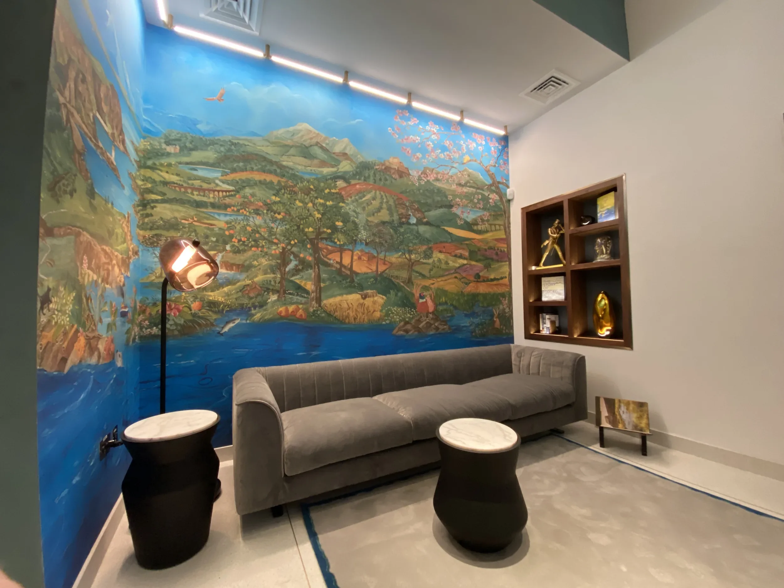 Interior of a room with a large colorful mural of mountains, trees, a river, and pink cherry blossoms on the wall. There is a gray sofa, several black and white side tables, a floor lamp, a wooden shelf with decorative items, and a small painting on an easel.