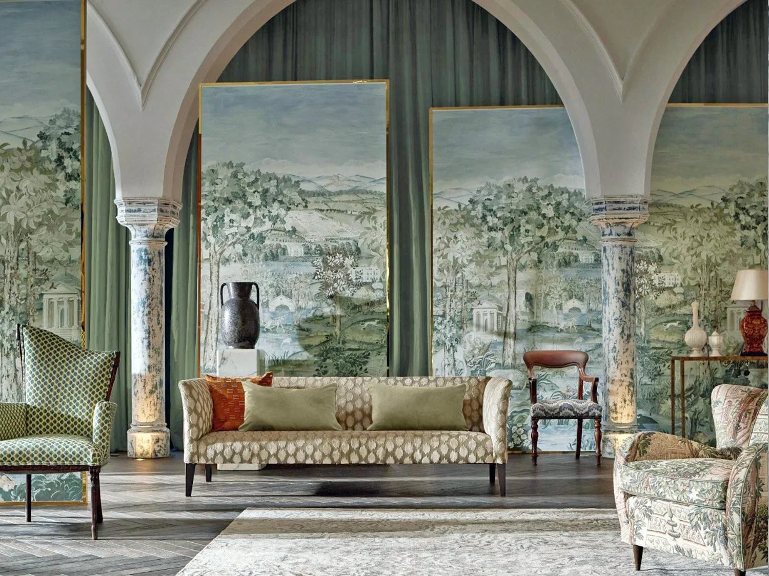 An elegant living room with a mural wall of scenic landscape, featuring trees, hills, and classical architecture, flanked by two large columns, with vintage furniture including a sofa, armchairs, and decorative items.