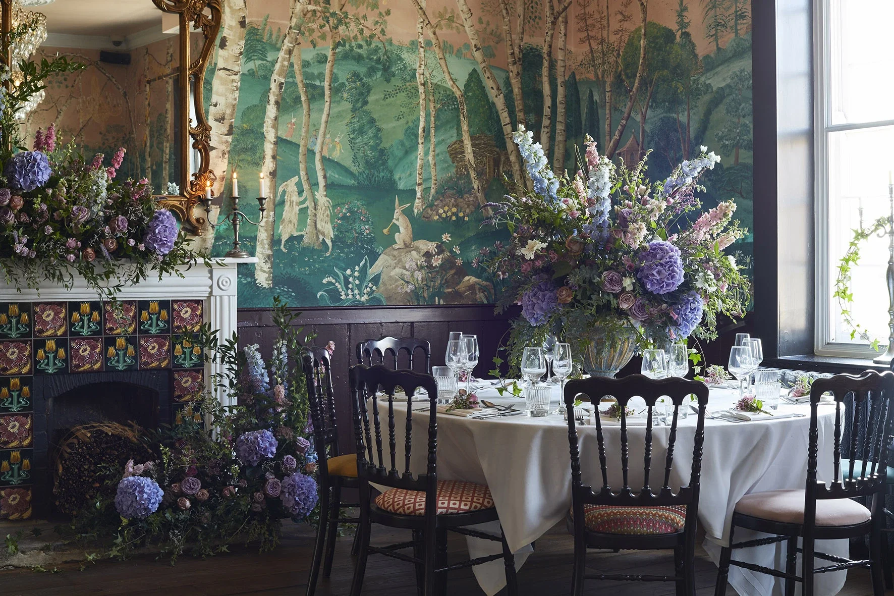 An elegant dining room with a table set for a formal meal, decorated with a large floral centerpiece of purple and white flowers. There is a large mural of a forest scene with trees, rabbits, and a fox in the background. The room has a window on the right, a fireplace with a floral arrangement on top, and a large ornate mirror above the fireplace.