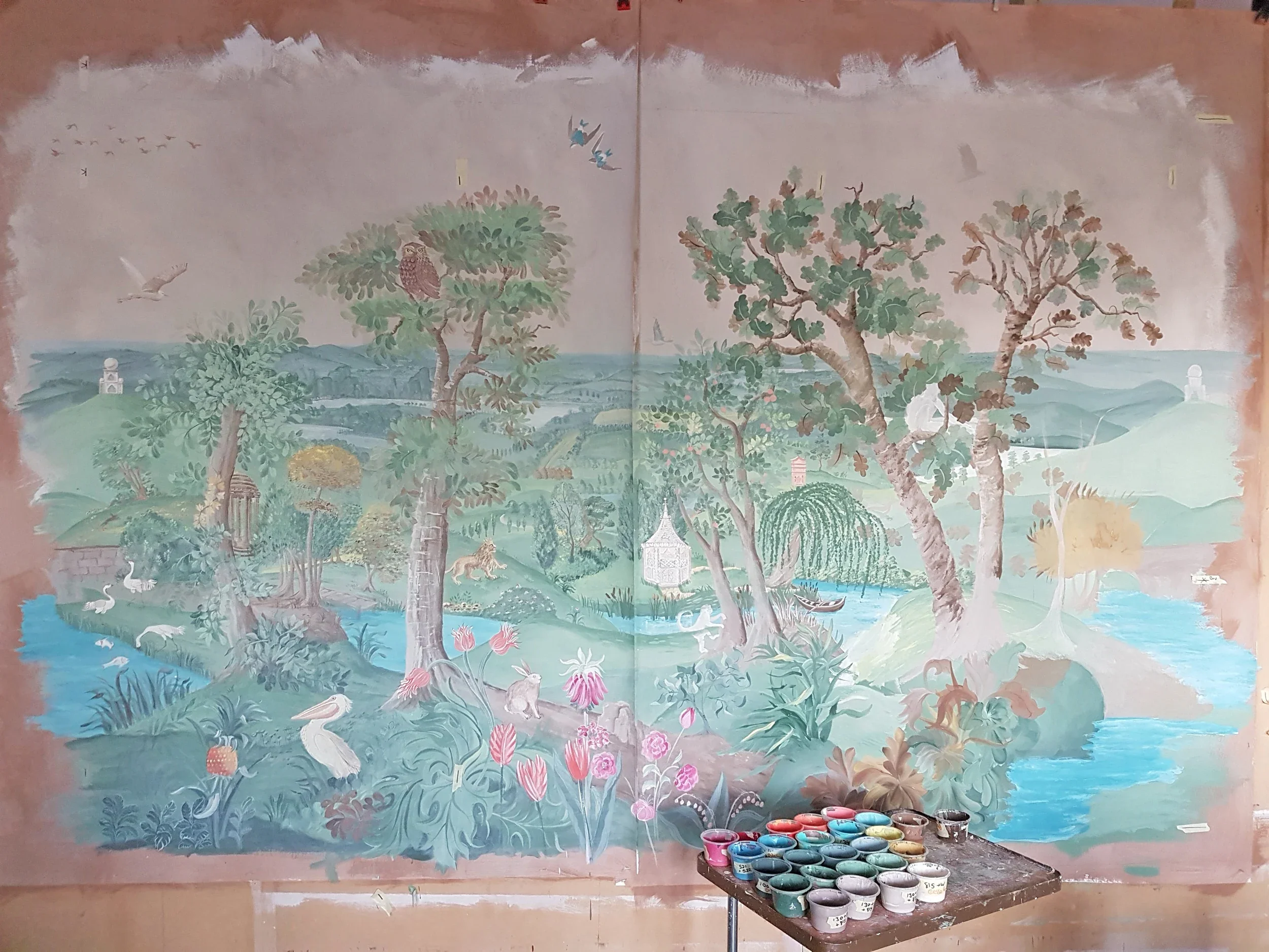A large traditional Asian landscape mural with trees, water, and animals, along with a palette of paint cans placed on a table.