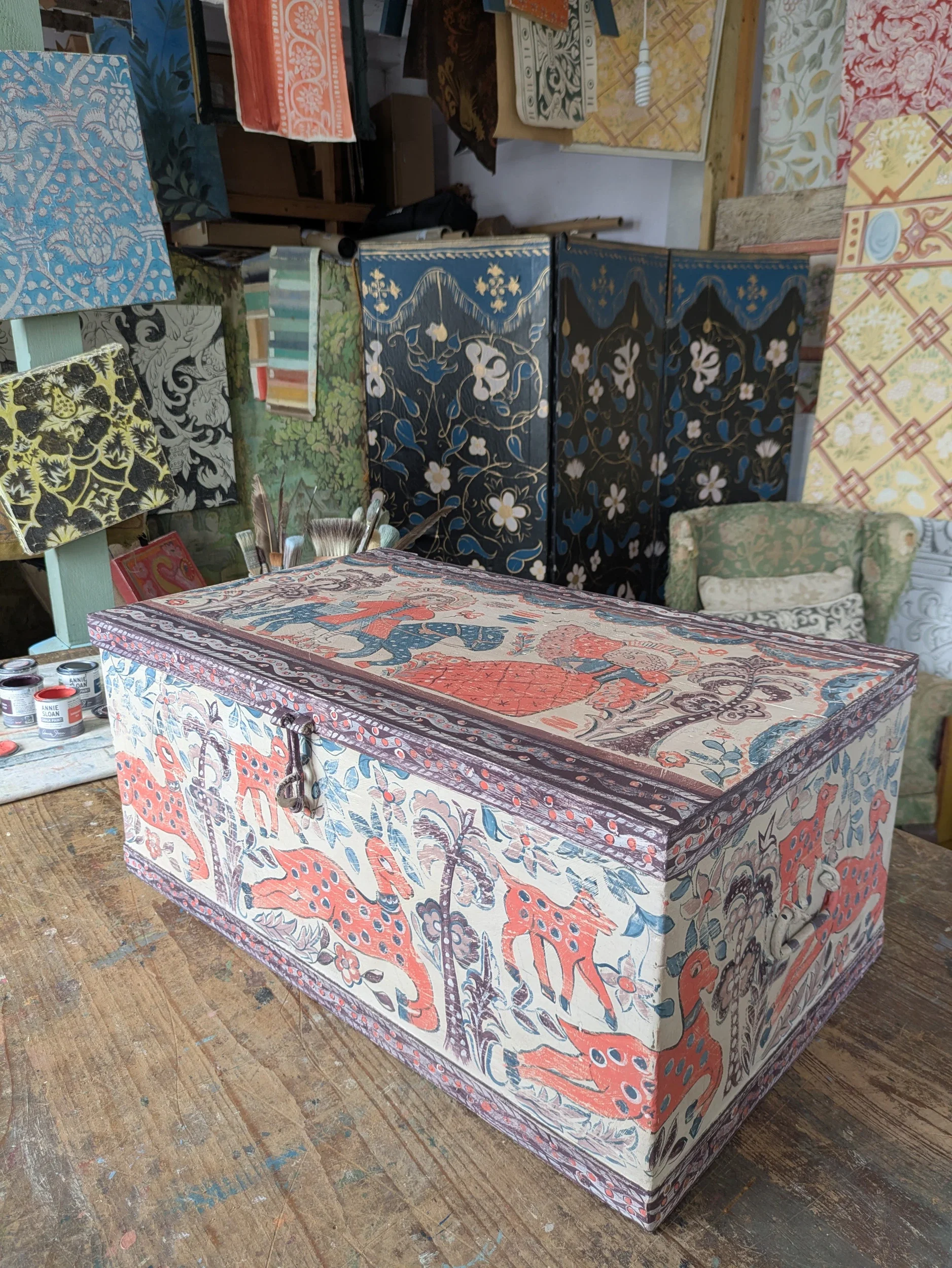 A collection of fabric-covered boxes and furniture pieces with various colorful patterns, including floral, geometric, and abstract designs, in a workshop or studio setting.