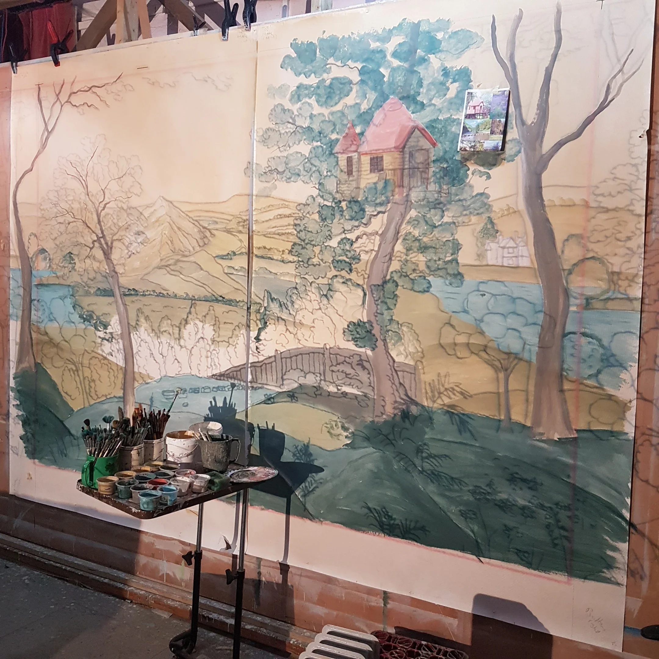 A large watercolor painting of a scenic landscape on a wooden panel, depicting trees, mountains, a house with a red roof, and a river, with artist supplies such as paint containers, brushes, and palettes in front.