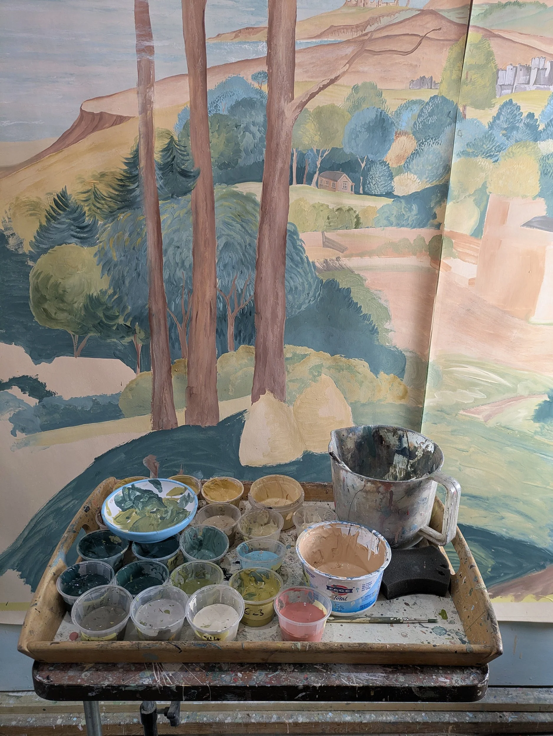 A painter's palette with small containers of various colors of paint, including blue, green, pink, and beige, is placed on a table in front of a partially painted landscape mural featuring trees, hills, and a small house.