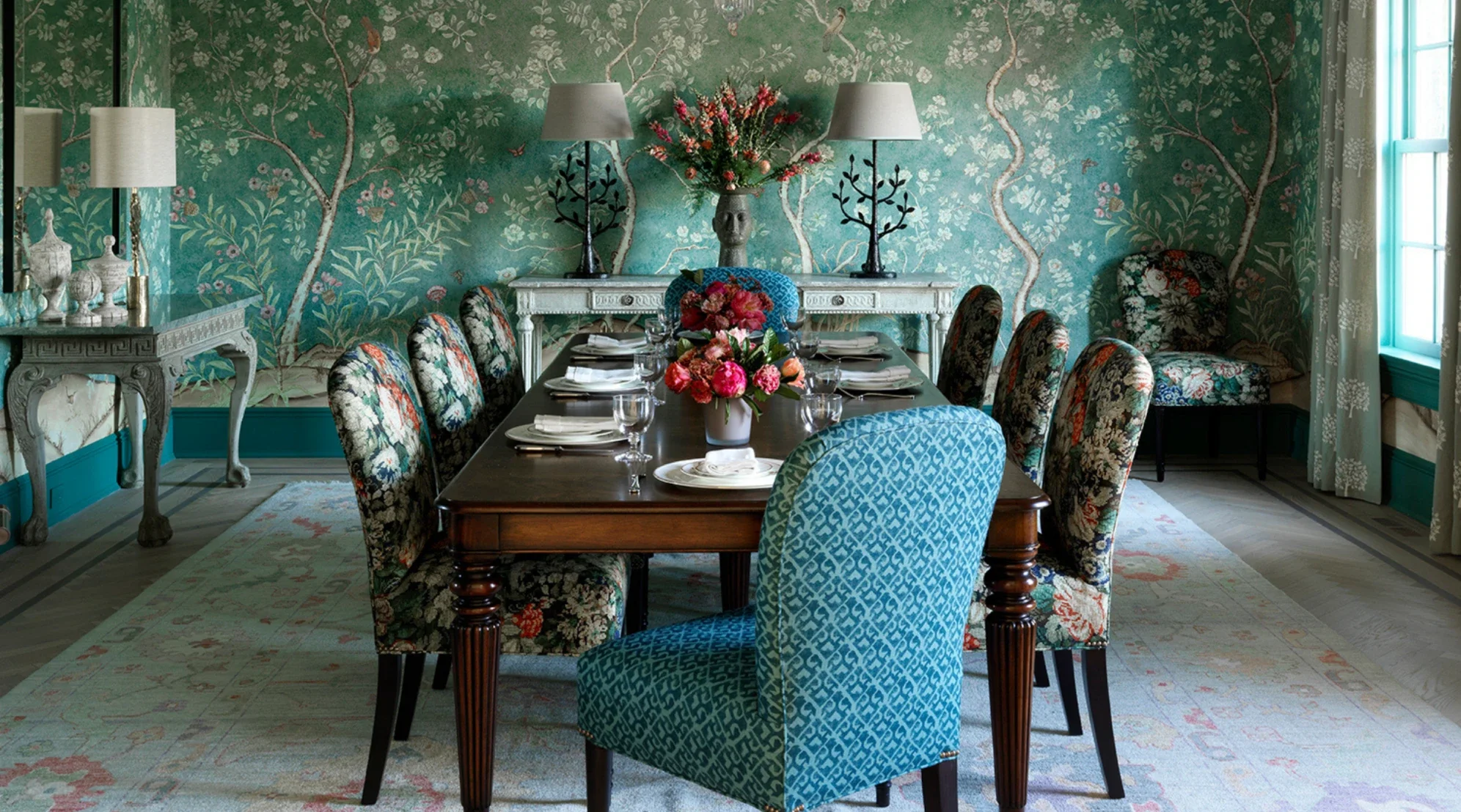 A dining room with a dark wooden table set with plates, glasses, and napkins. There are eight upholstered chairs with floral and geometric patterns around the table. The room features teal walls with a floral mural, and a rug with floral designs covers the floor. Two white table lamps, a large floral arrangement, and decorative objects are on side tables against the wall. A window with white curtains is on the right side of the room.