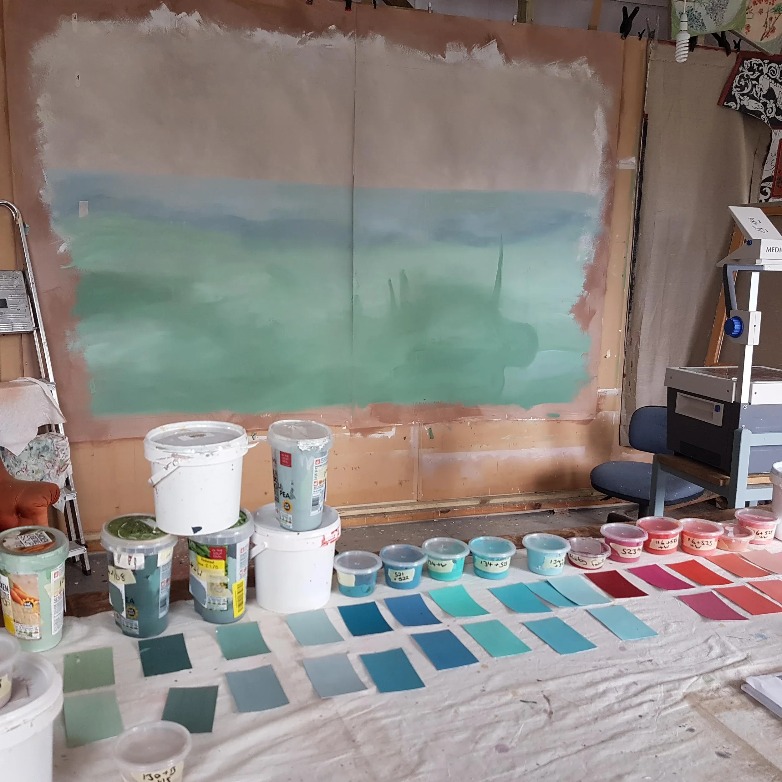 A painting in progress of a landscape on a large canvas, with paint buckets, color swatches, and painting supplies scattered on the floor and table nearby.