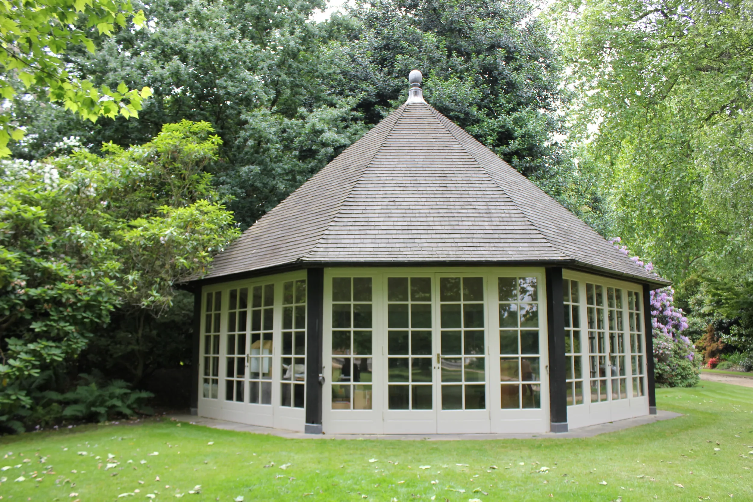 A round gazebo with large windows and a shingled, conical roof, surrounded by lush green trees and a neatly trimmed lawn.