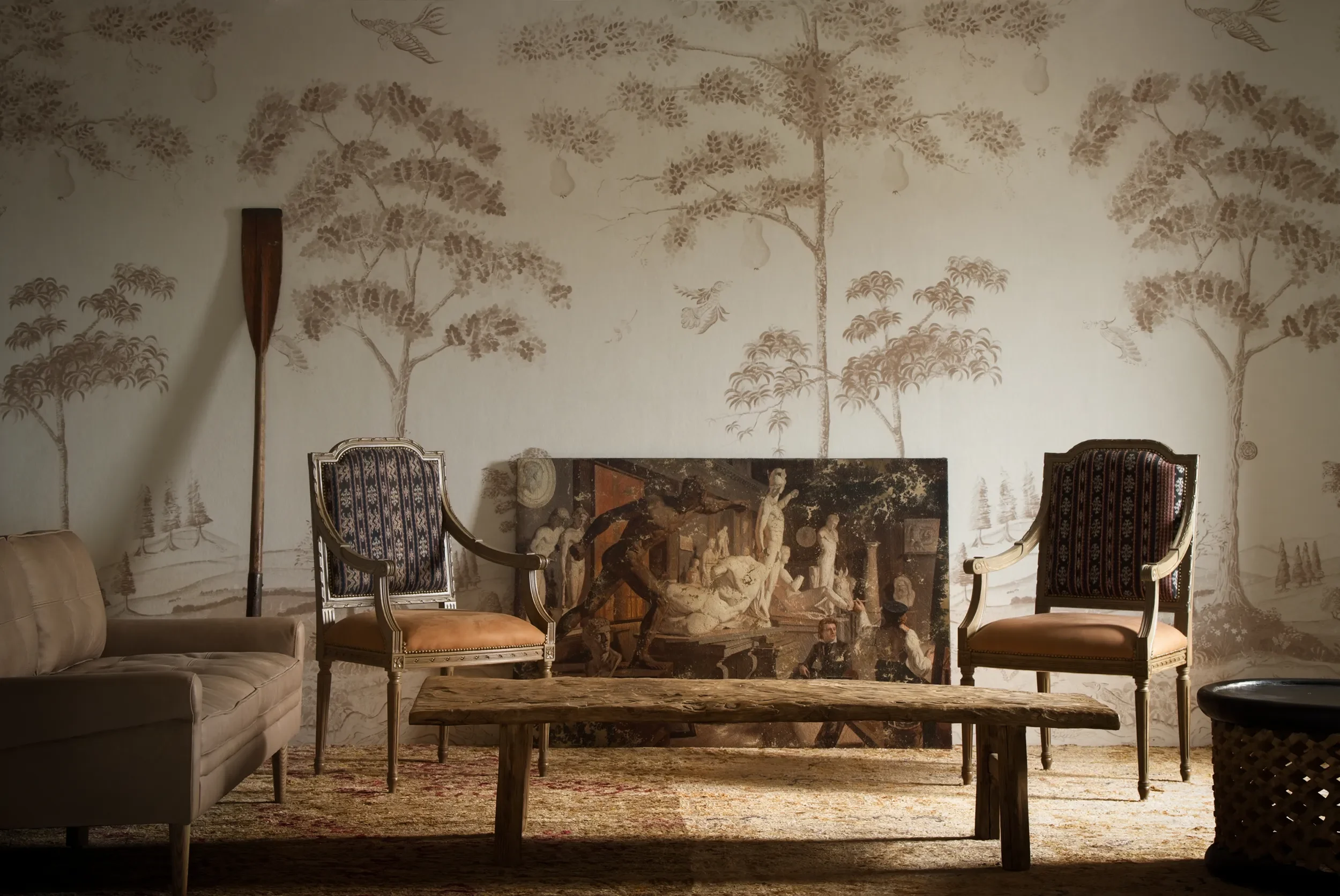 Melissa White's Mythical Land wallpaper in a more neutral colour-way. Wallpaper is with Kit Kemp for Andrew Martin. Scene shows antique wooden furniture with an oil propped against the wall.