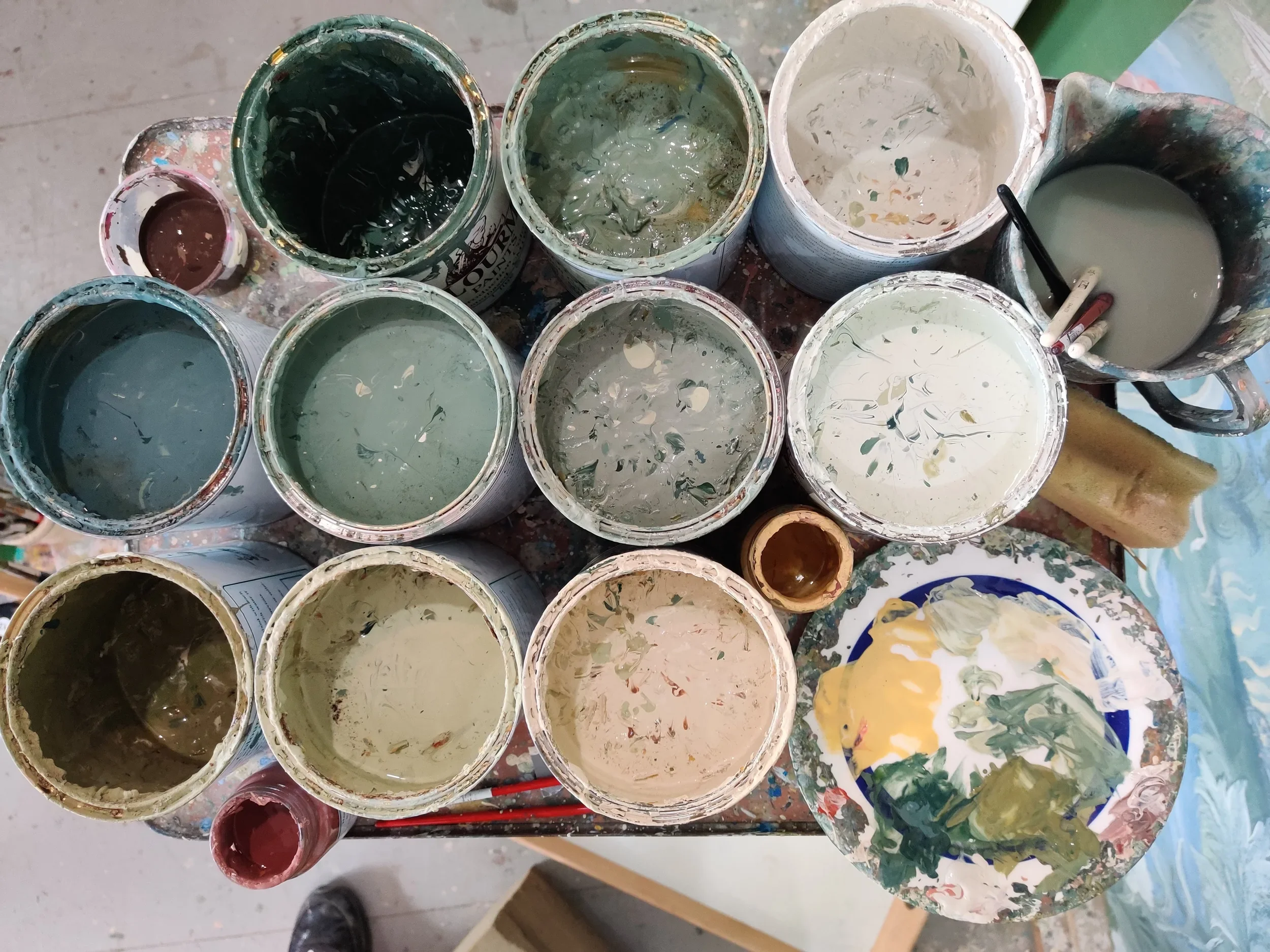 View of multiple open paint cans with various shades of green, beige, and red paint, some with spilled paint around the rims, placed on a splattered work surface, and a plate with paint marks nearby.