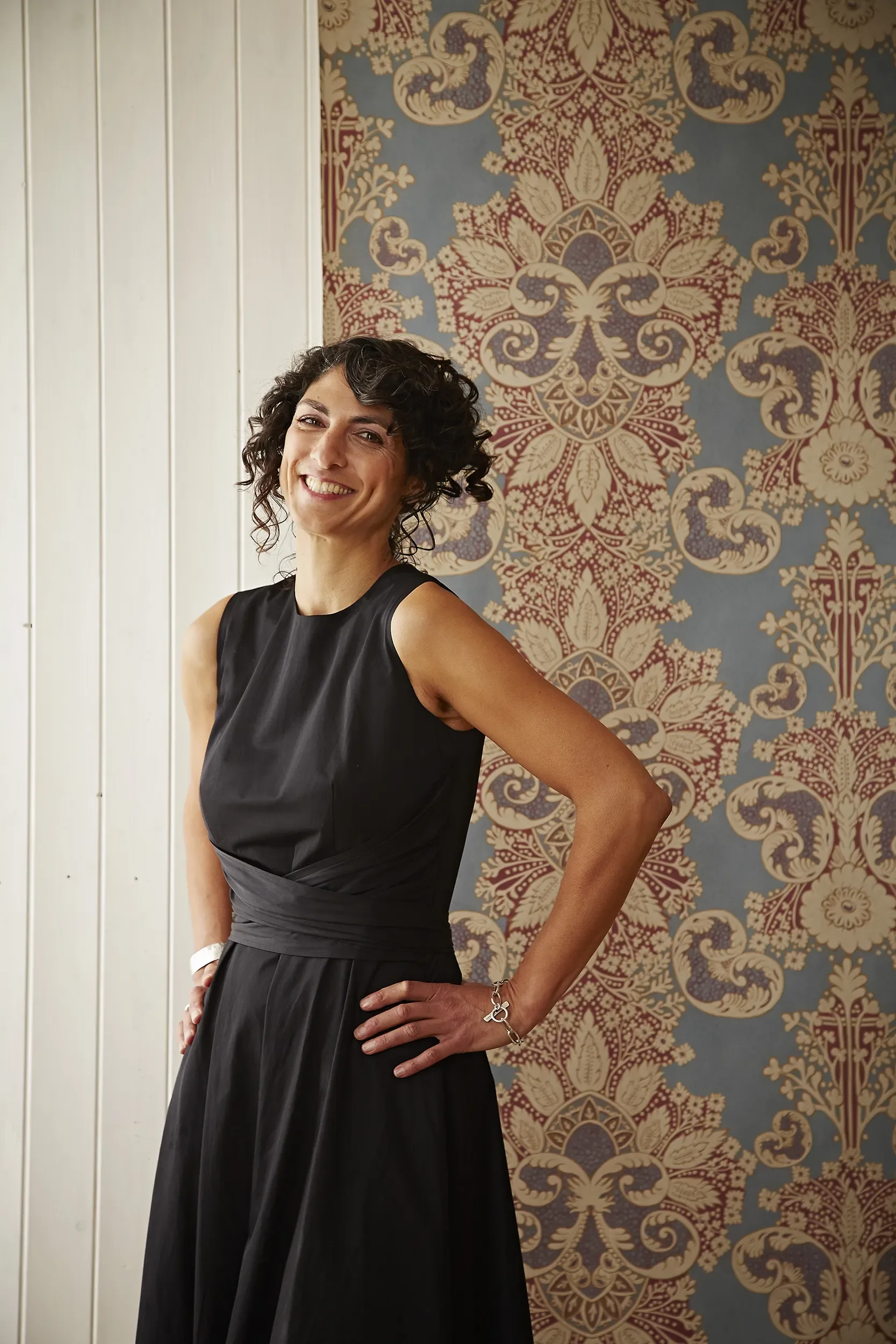 Melissa White is pictured hands on hips in a blue sleeveless dress in front of her Rococo wallpaper for Lewis & Wood.