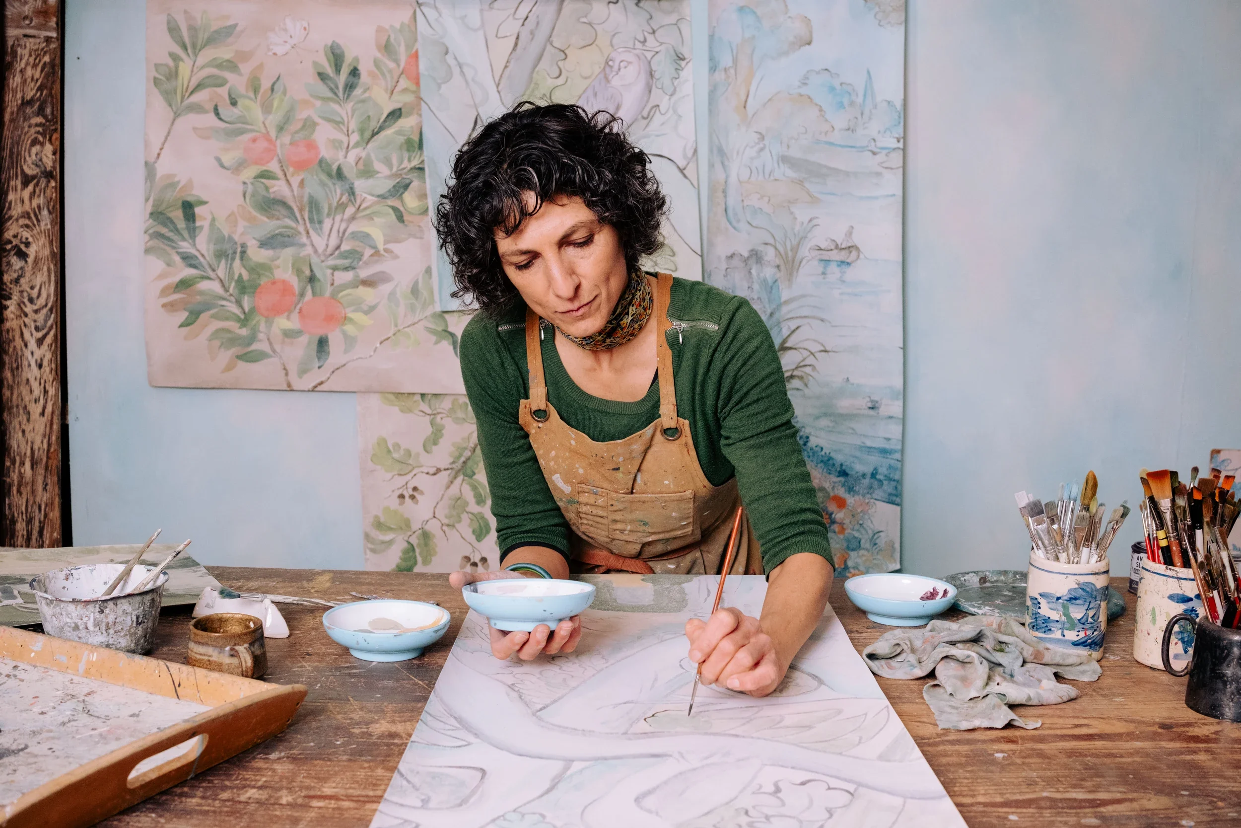 A woman with dark curly hair wearing a green shirt and apron painting a large sheet of paper with watercolors at an art studio. There are bowls of paint and brushes on the table, and watercolor paintings of nature scenes hang on the wall behind her.