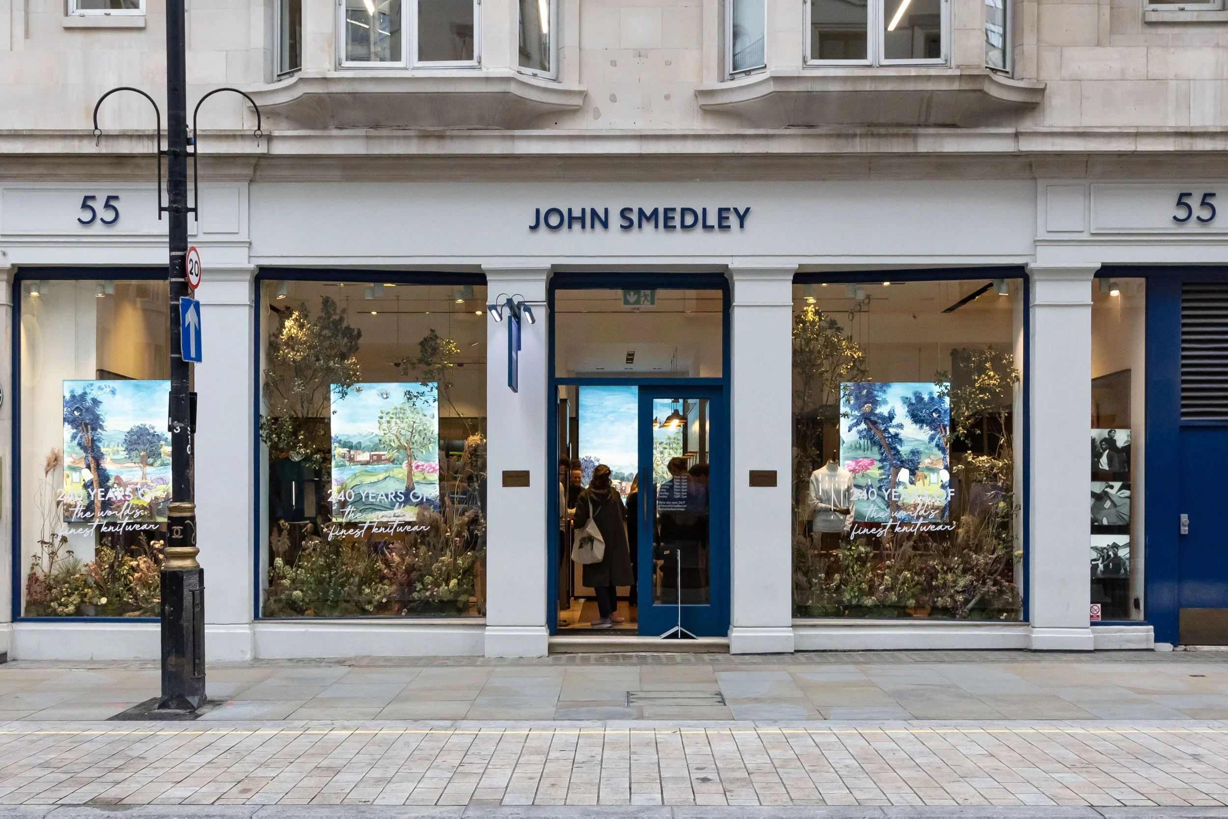A storefront with the name John Smedley, displaying large store windows with mannequins and promotional posters. The street in front has a lamppost and sidewalk tiles.