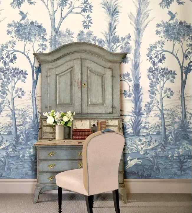 A vintage dresser with a weathered blue finish, topped with a bouquet of white and green flowers in a beige vase, positioned in front of a decorative blue and white wallpaper with nature scenes, and a beige upholstered chair with dark wooden legs in the foreground.
