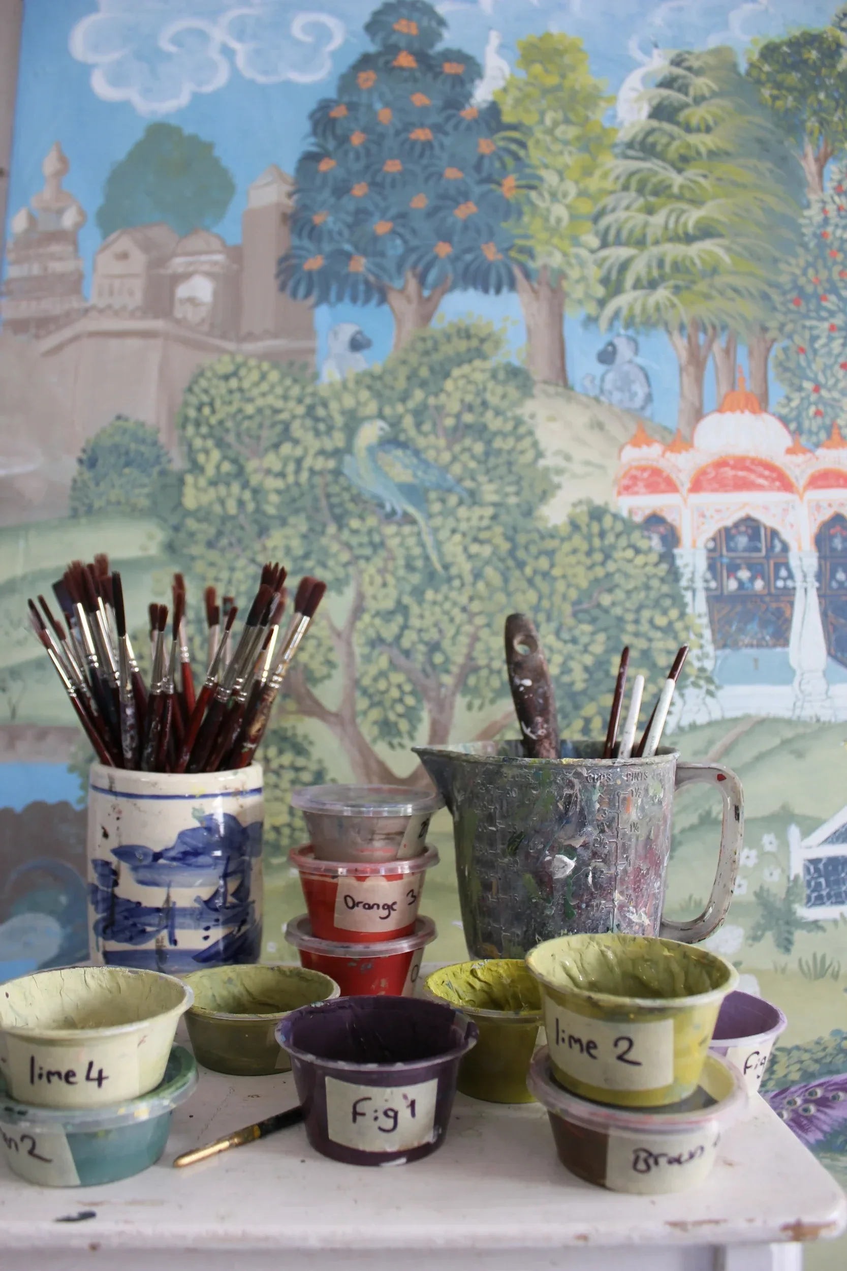Painting supplies including paintbrushes and various containers of paint labeled with colors such as lime, orange, purple, and brown, set in front of a colorful mural featuring trees, clouds, and architectural structures.