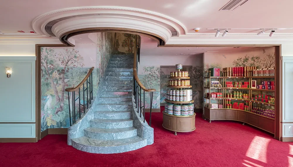 Indoor space with a curved stone staircase, ornate wallpaper with nature scene, red carpet, bookshelf with colorful books and decorative tins.
