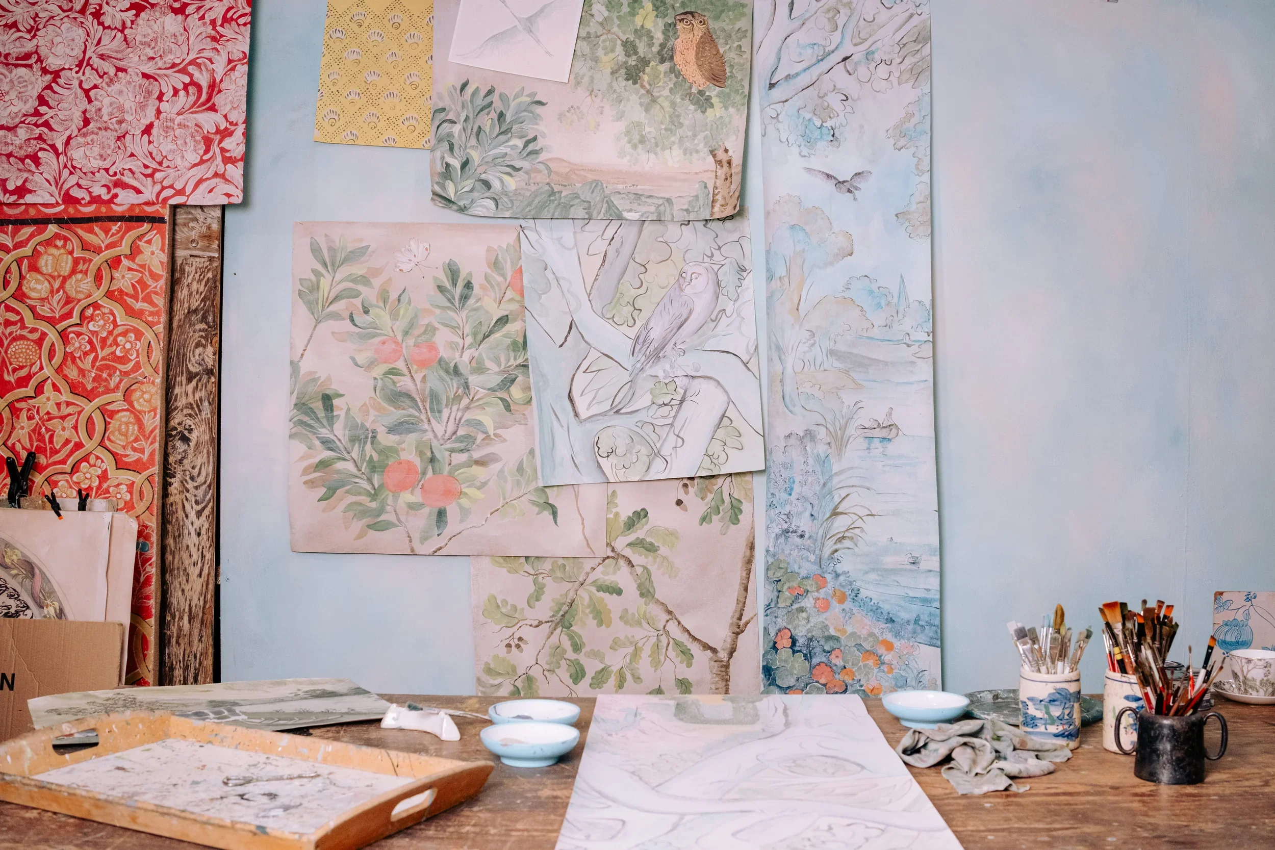 A workspace with various watercolor paintings of nature scenes hung on a light blue wall, an art table with paper, brushes, and bowls of water in the foreground.