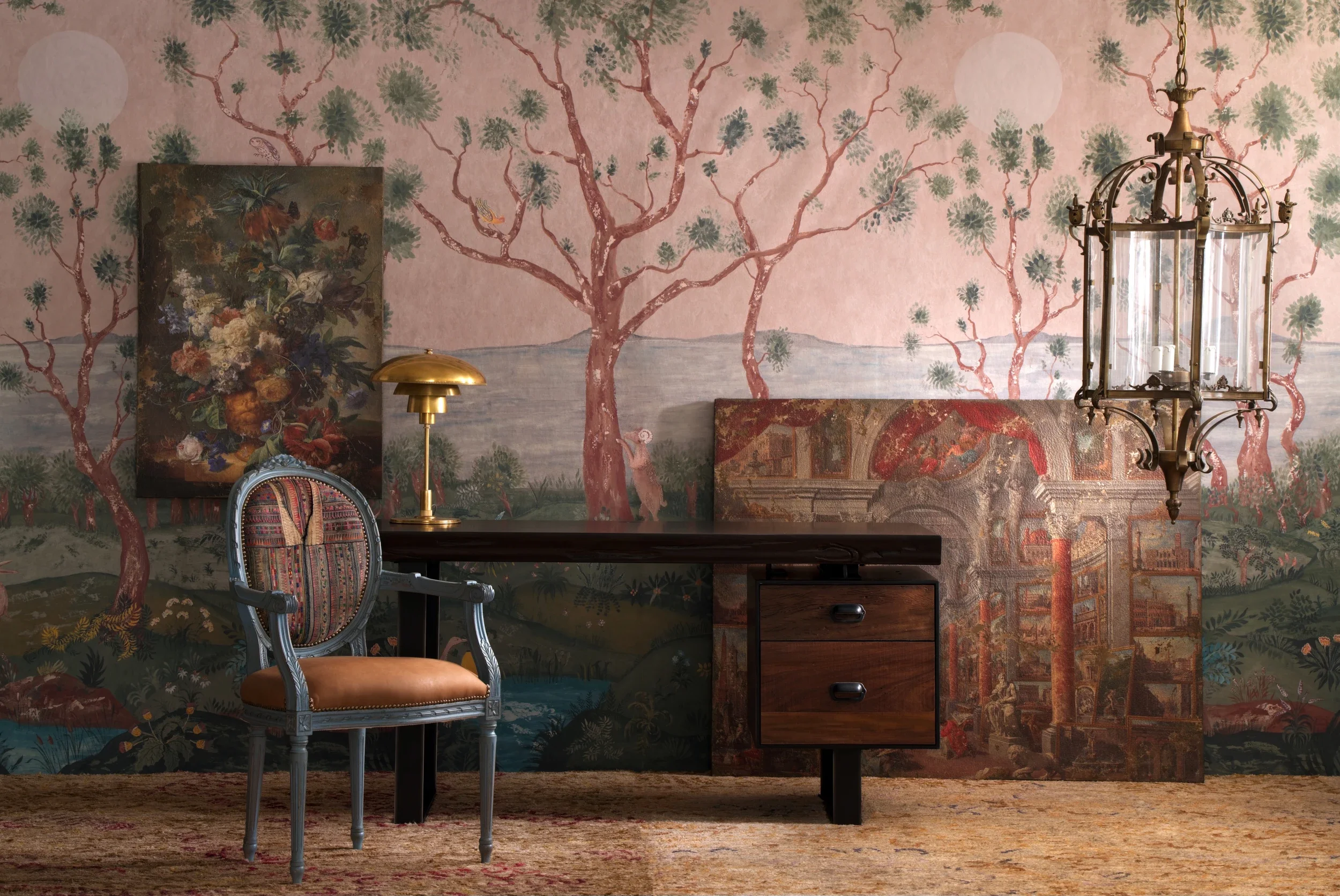 Interior of a room with a painted wallpaper mural of trees, water, and nature scenes. There is a vintage armchair with a patterned fabric, a wooden dresser with three drawers, a brass desk lamp, a large painting of flowers, and a decorative brass and glass chandelier.