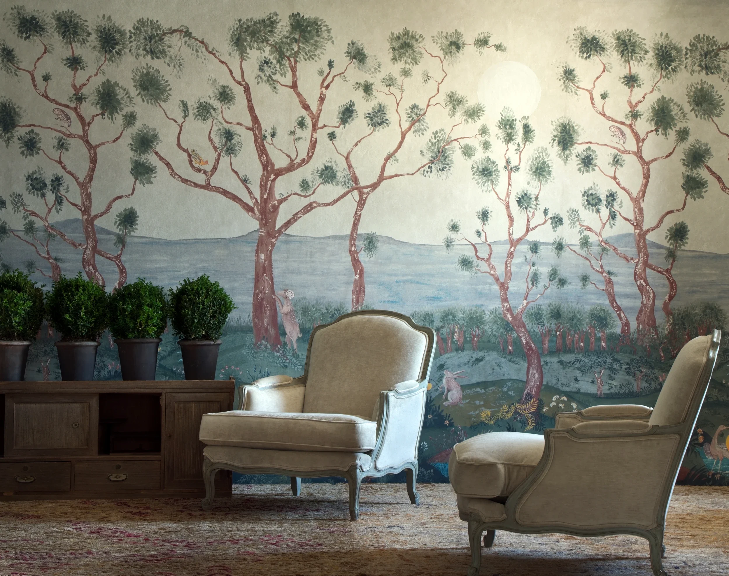 A mural painting of trees, animals, and a landscape scene on a wall with white upholstered armchairs and a wooden cabinet in front.