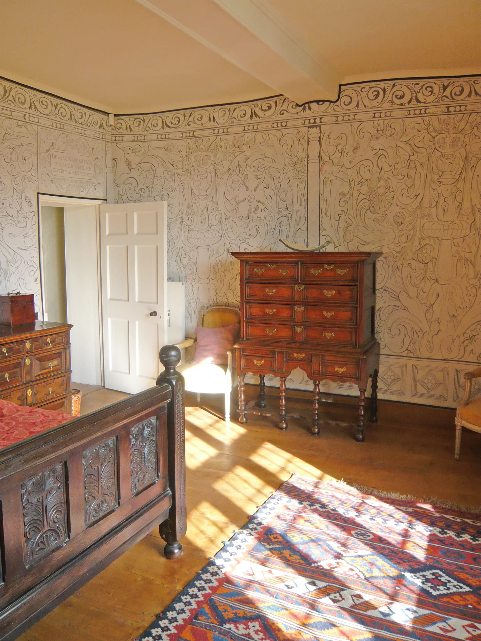A room with wooden furniture, including a carved wooden bed, a tall dresser, and a chair with a pillow. The walls are decorated with intricate line drawings and Latin text. A colorful, patterned rug is on the wooden floor, and sunlight is shining into the room.