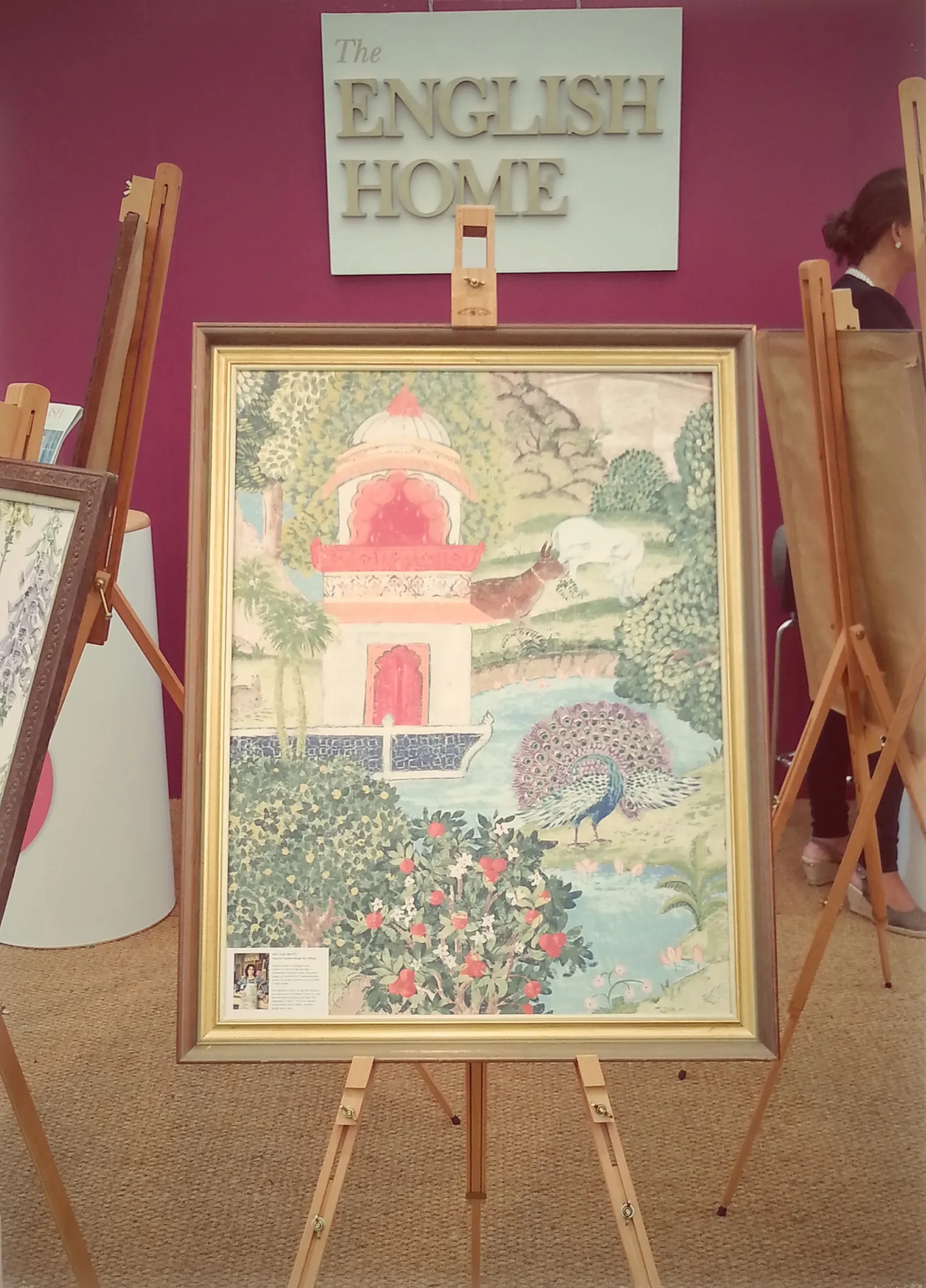 An art gallery display features a framed painting of a traditional Asian scene with trees, a pavilion, and a peacock in a garden. The painting is positioned on an easel, with a large sign above that reads 'The English Home' on a purple wall.