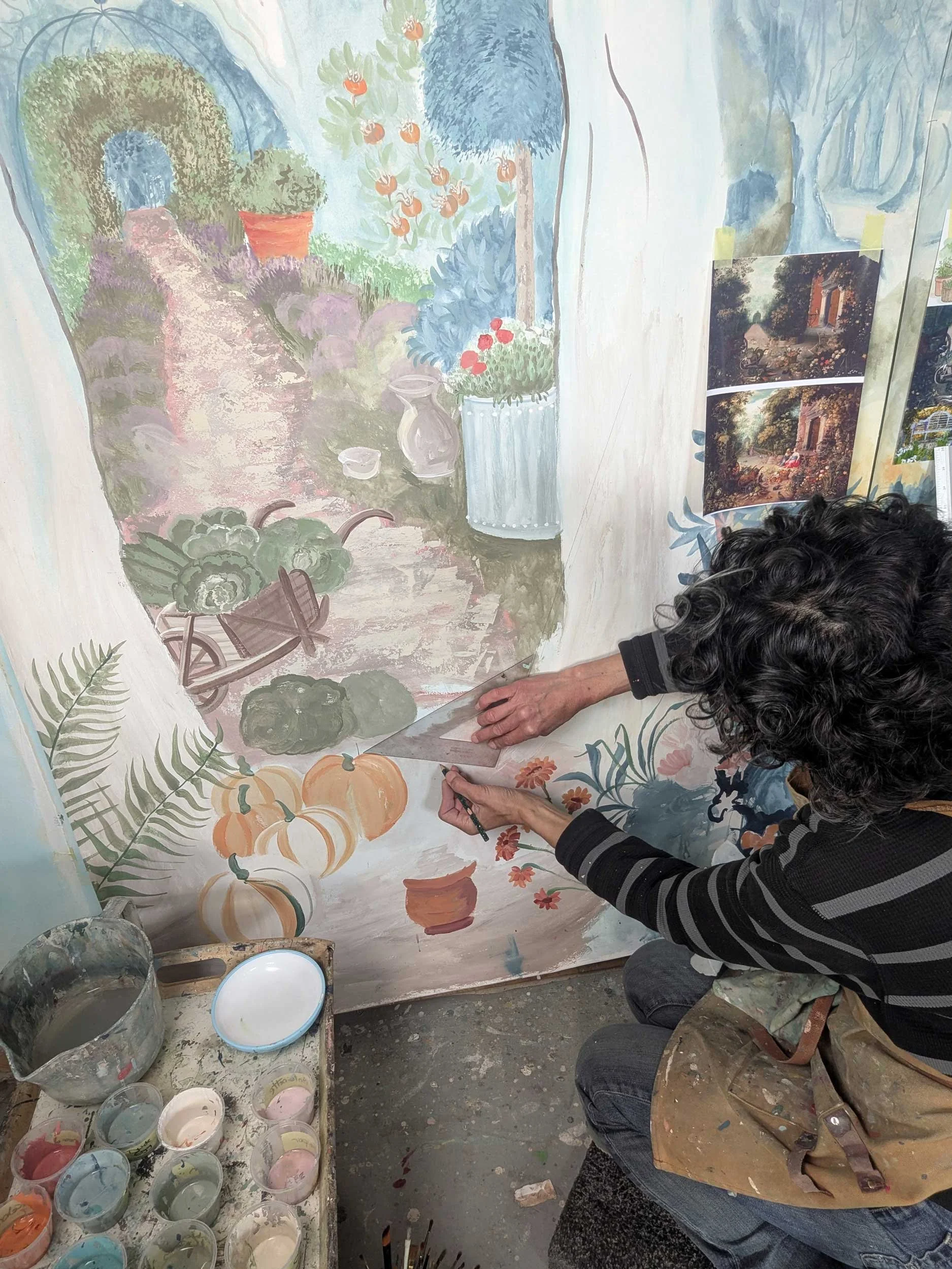 Melissa White painting the detail of a garden rake leaning against a tree on the full-scale mural artwork.