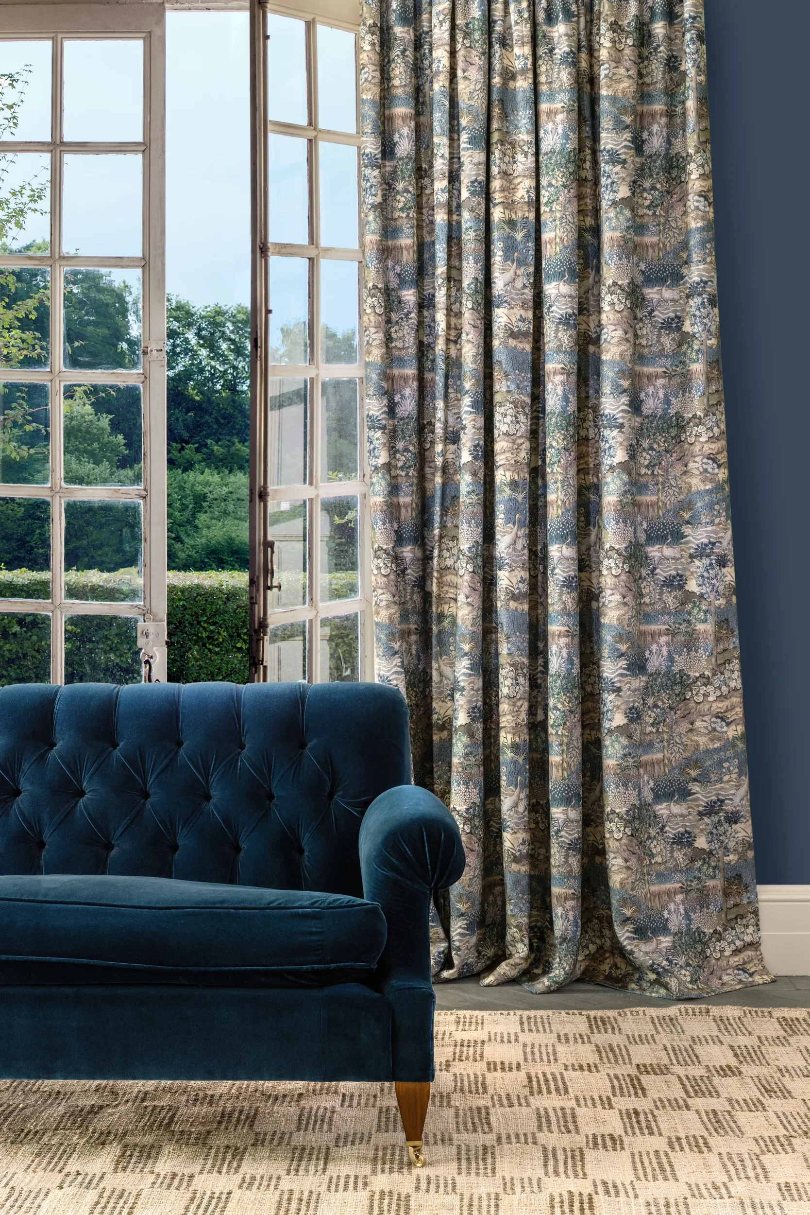 A living room with a large window and a long, floral patterned curtain. Part of a blue velvet tufted sofa and a beige patterned rug are visible.