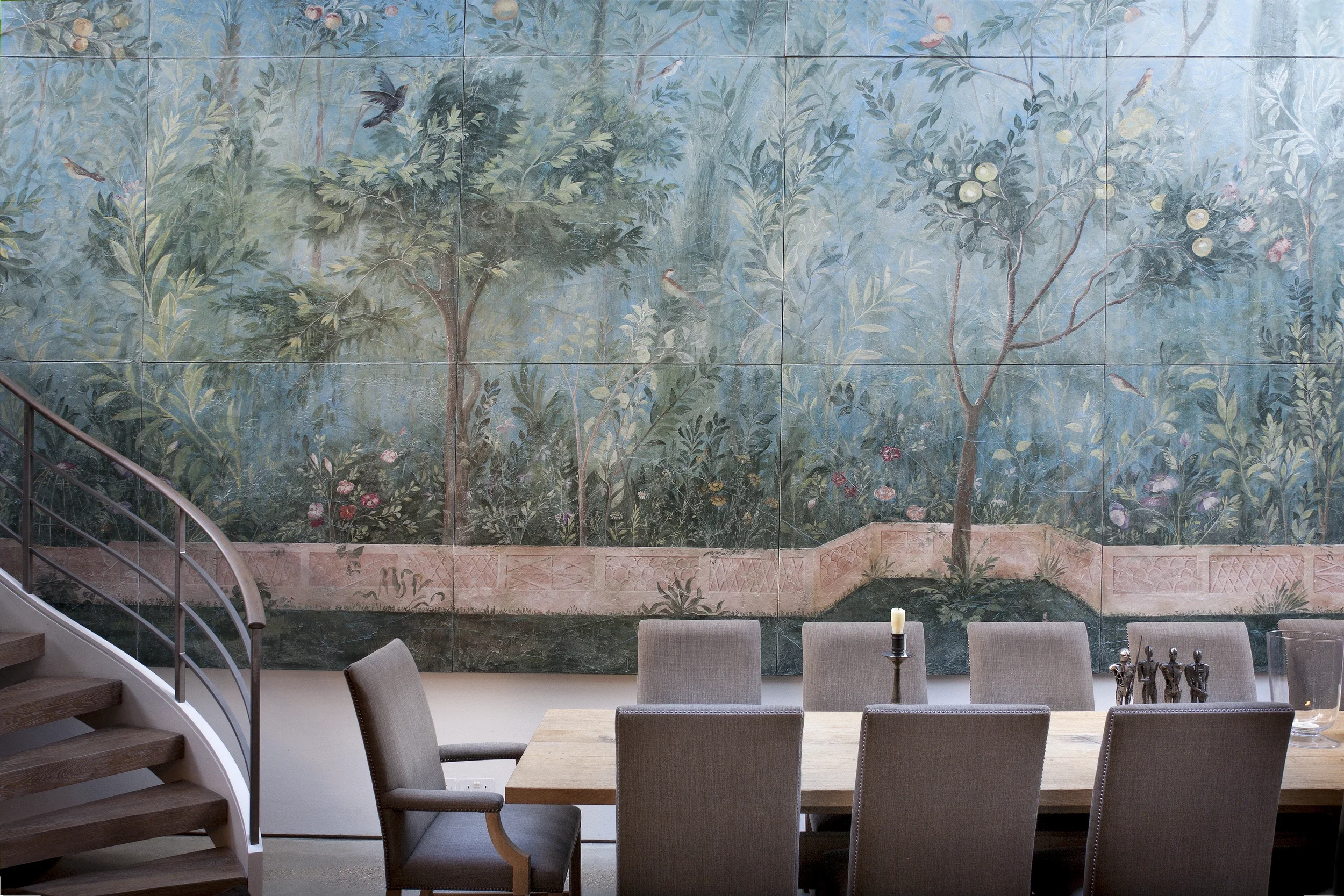 A large wall mural depicting a lush garden scene with trees, birds, and fruit, located in a dining or conference room, partially visible stairway on the left, chairs around a table in the foreground.