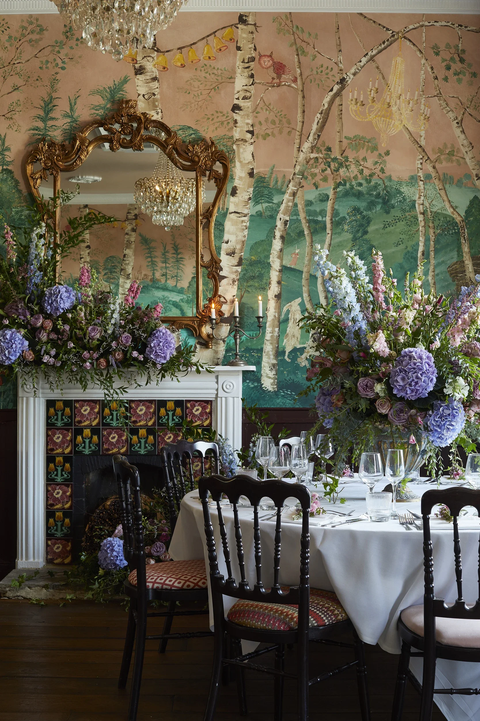 The Big Room at The Bell in Ticehurst, East Sussex, showing the bespoke scenic wallpaper by Melissa White, 2020. Hand-painted woodland landscape with silver birch trees, table set for dining with large floral arrangement and decorated fireplace.