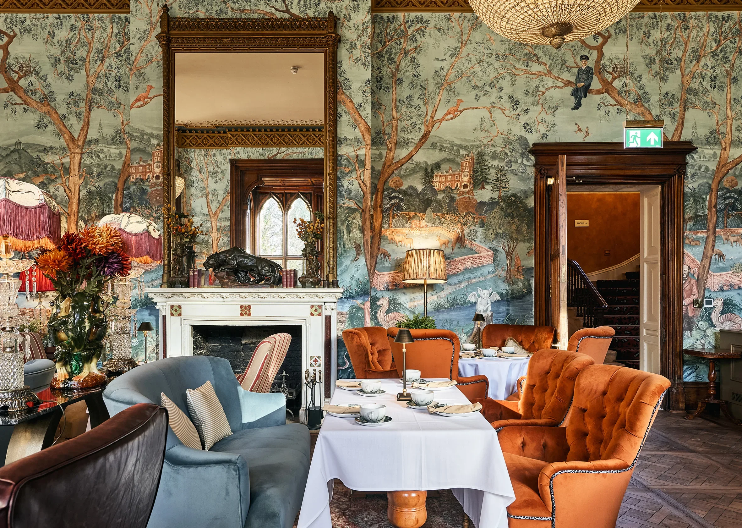 Elegant dining room with colorful tufted chairs around a table set with white tablecloths and fine china. Ornate woodwork, a marble fireplace with floral arrangements, a large landscape mural with trees, animals, and buildings, and vintage decorative lamps.