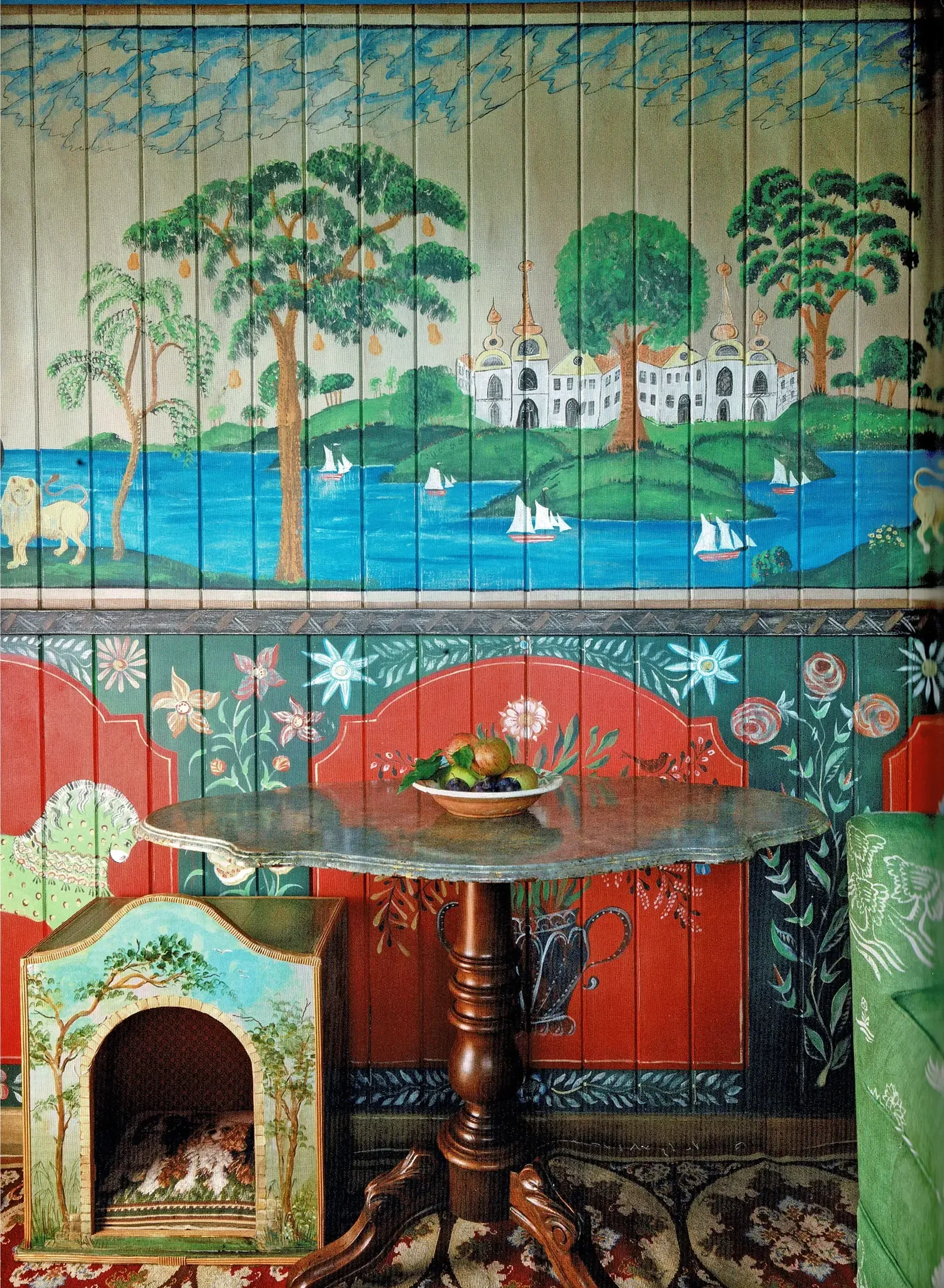 Decorative wall mural with painted landscape featuring trees, water with sailboats, and a building with golden domes. There is a wooden table with a bowl of fruit in front of the mural.