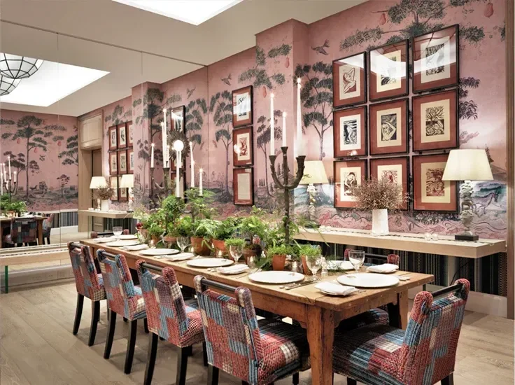 A dining room with a decorated wall featuring framed artwork and a mural of trees, a wooden table set with plates and glasses, colorful upholstered chairs, and multiple candelabra-style chandeliers with lit candles.