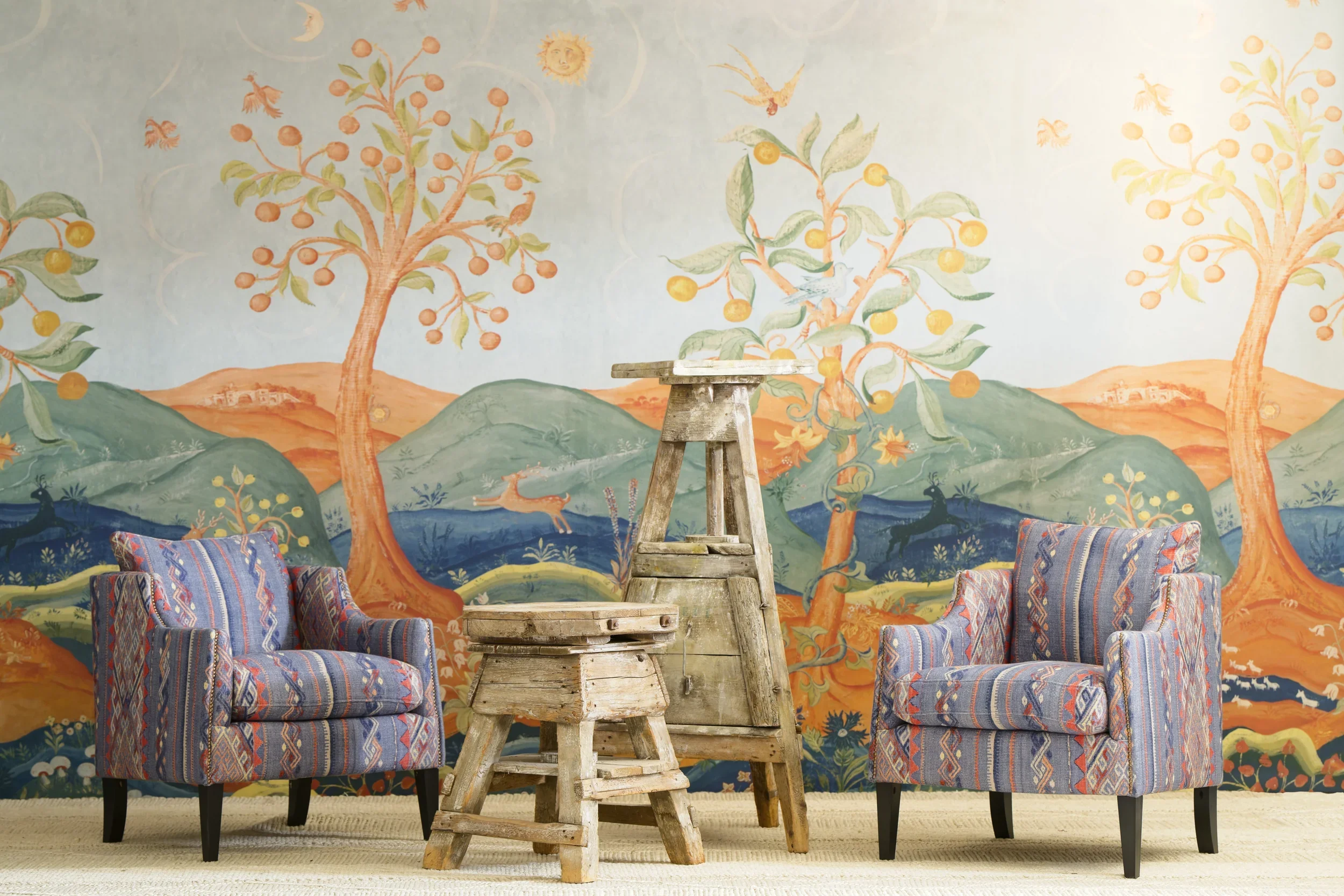 A decorative wall mural of trees and animals, with a central wooden stool, and two colorful patterned chairs in front.