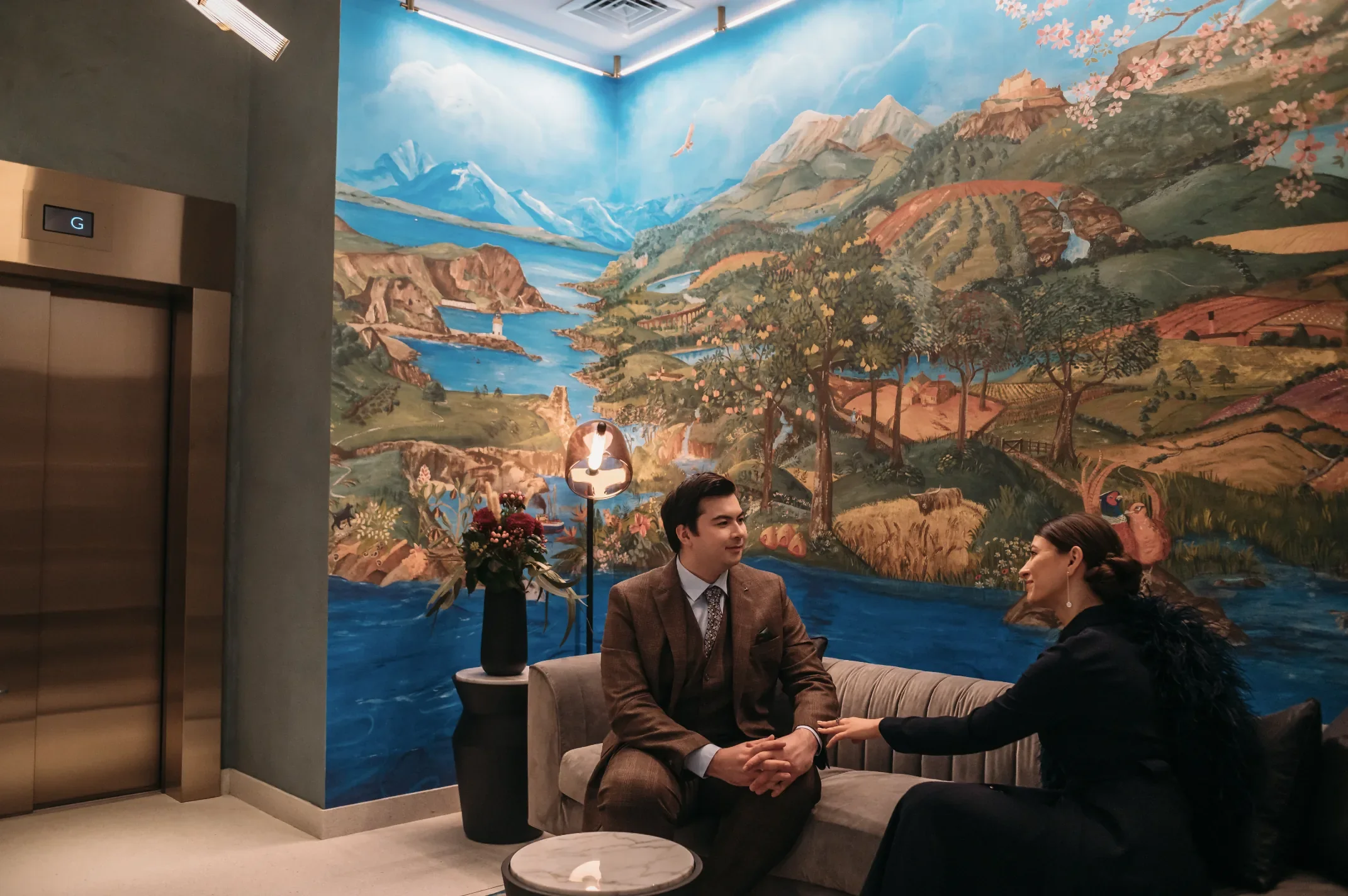 A mural depicting a scenic landscape with mountains, rivers, trees, and a cherry blossom tree on the right side. Two people are sitting on a sofa, holding hands and engaging in conversation. The scene is set in an indoor space, possibly a lobby or waiting area with sophisticated decor.